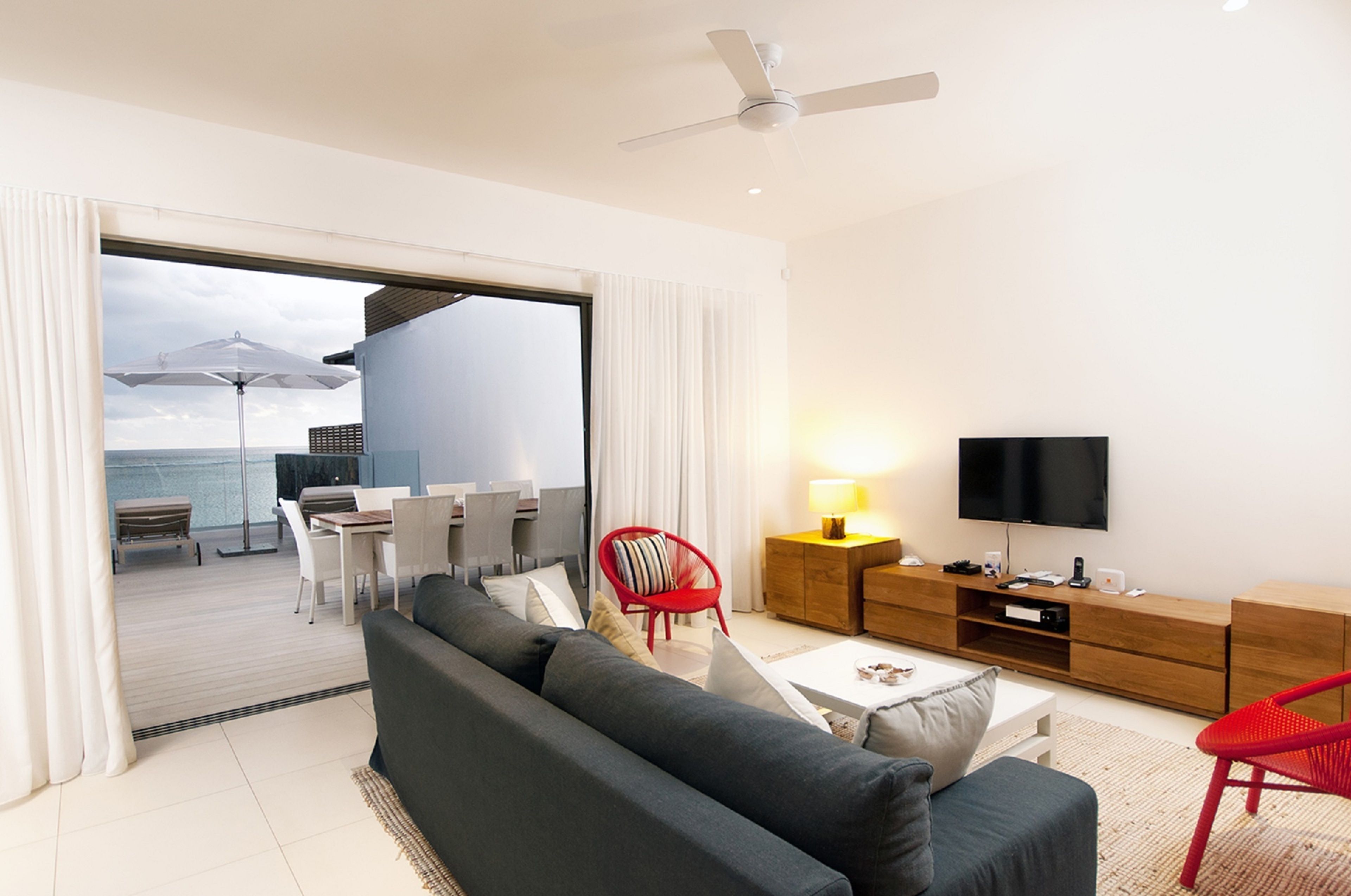 A modern living room with a sofa, red chairs, a TV, and a balcony with a view of the sea.