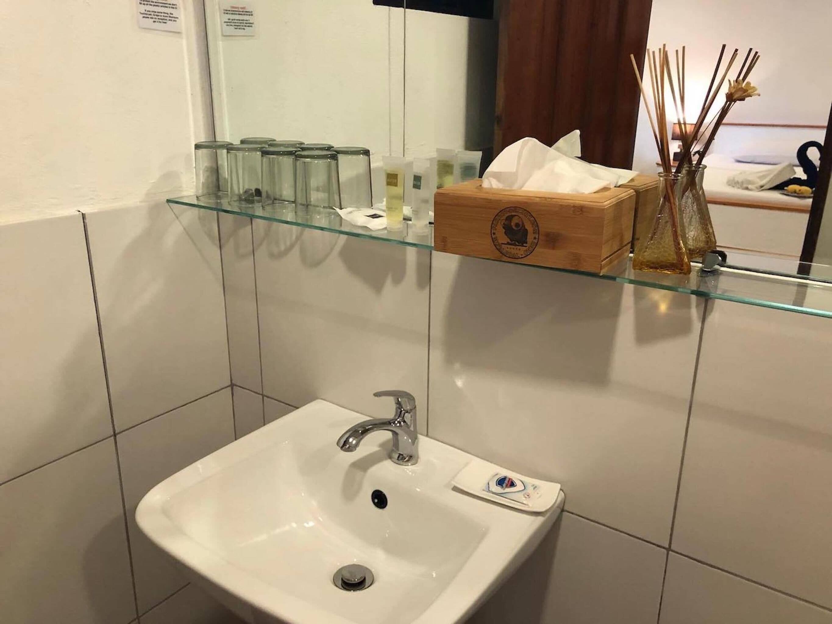 A bathroom with a white sink, glass shelves above containing toiletries, and a wooden box.
