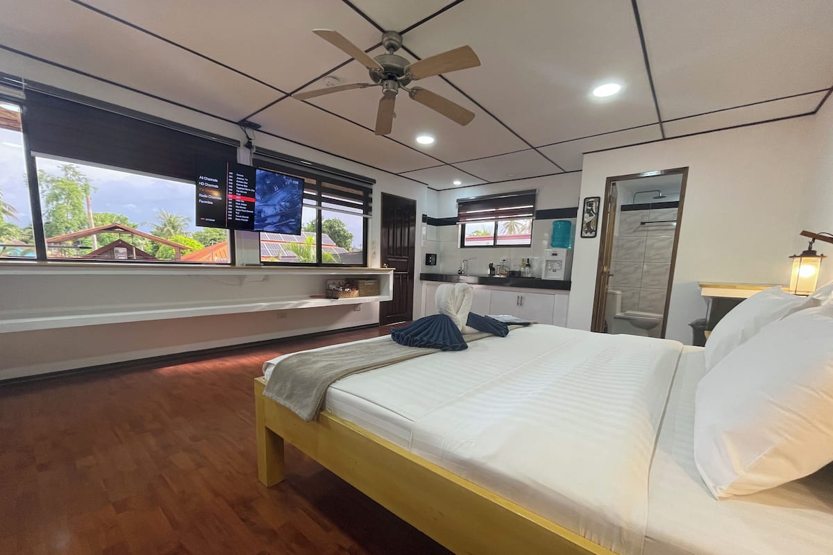 Quarto de hotel com uma cama, ventilador de teto, televisão, janela com vista para uma casa e banheiro com espelho e pia.