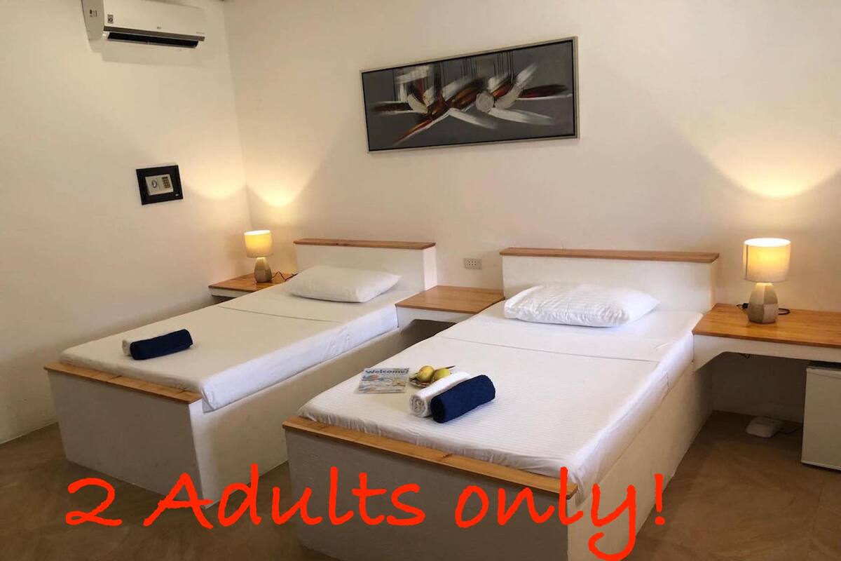 Quarto de hotel com duas camas de solteiro, cada uma com cabeceira de madeira, roupa de cama branca e uma pequena mesa de cabeceira com abajur. Um quadro está pendurado na parede acima das camas. O texto "2 Adultos apenas!" é visível no chão.