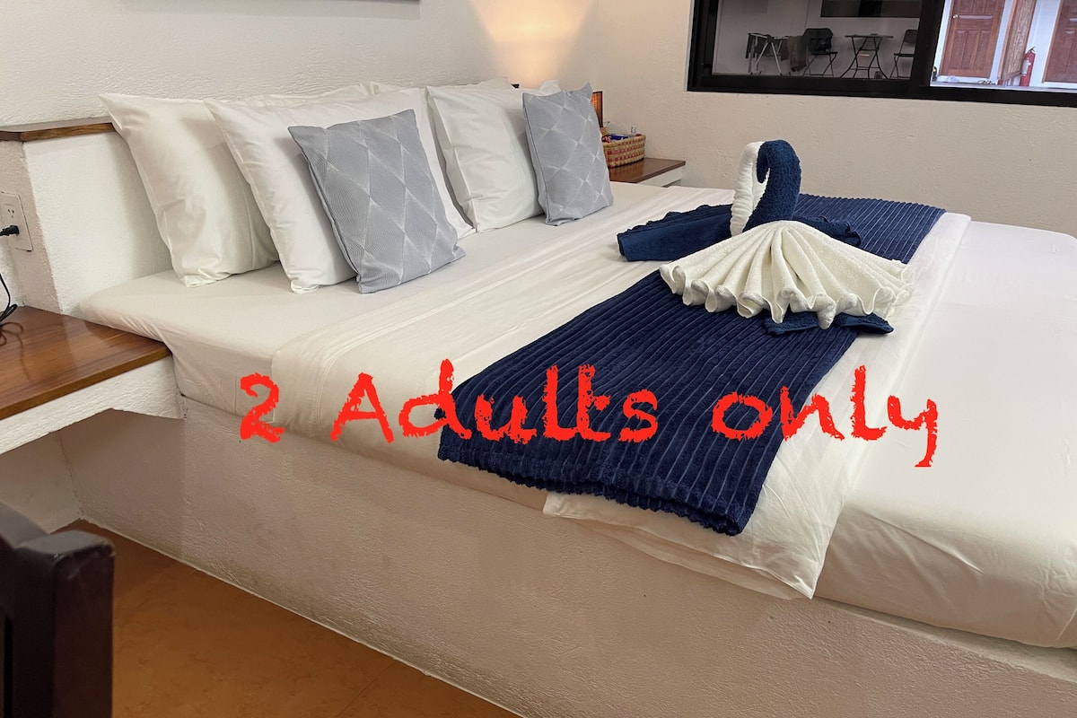 Uma cama bem arrumada com um cobertor azul e branco, travesseiros brancos e uma pequena mesinha de cabeceira ao lado.