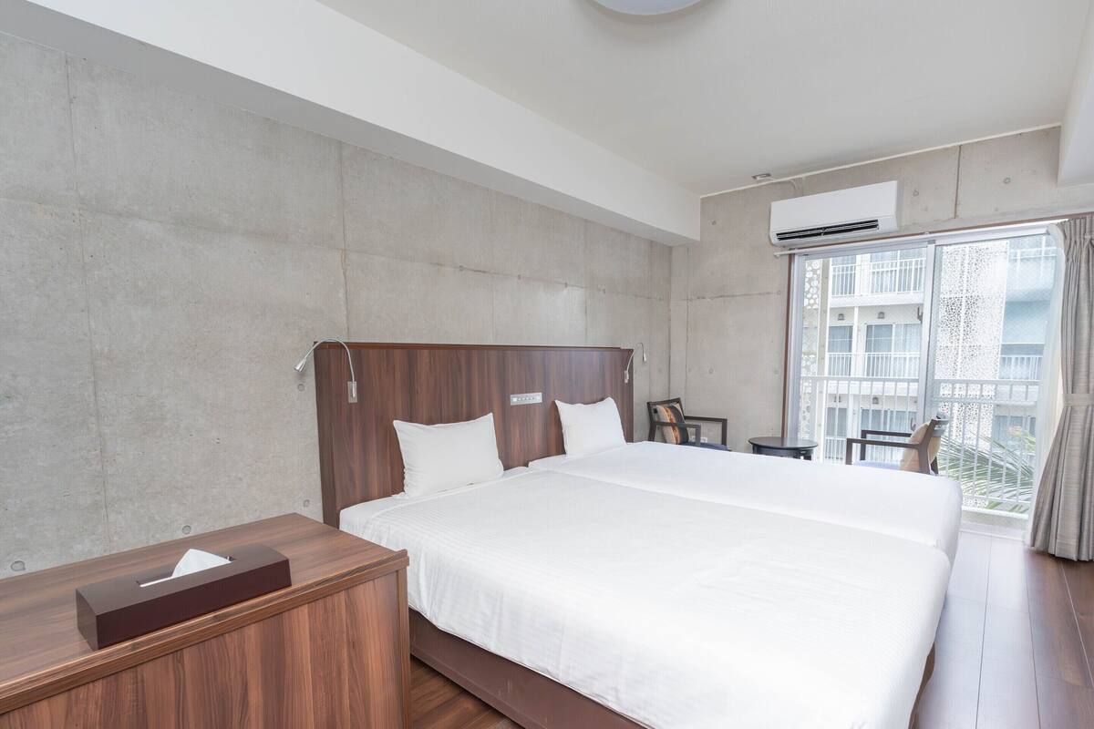 A hotel room with a large bed, a wooden bedside table, a desk with a chair, and a window with a view of buildings.