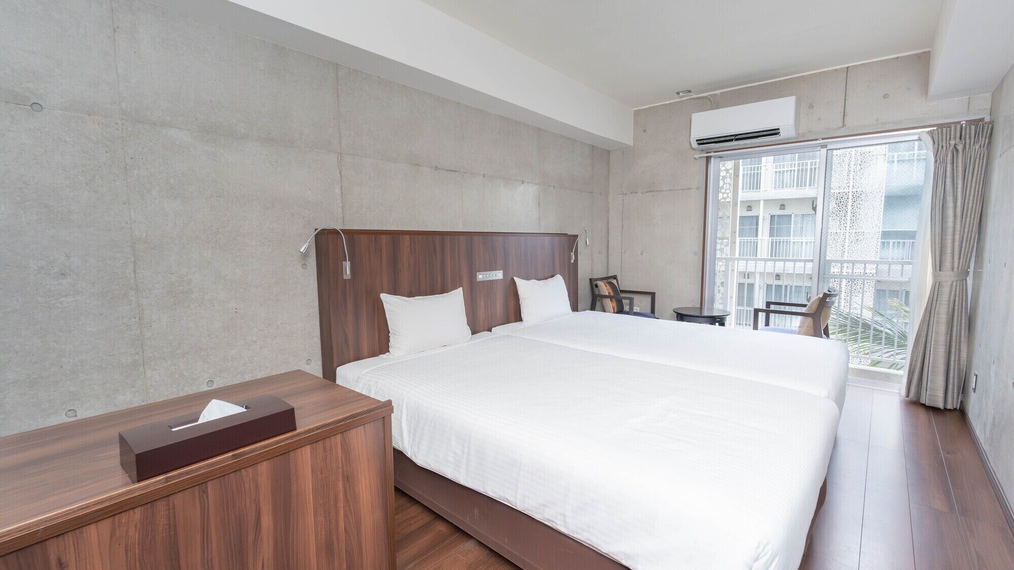 Habitación de hotel con una cama grande, una mesita de noche de madera, un escritorio y una ventana con vistas a un edificio.