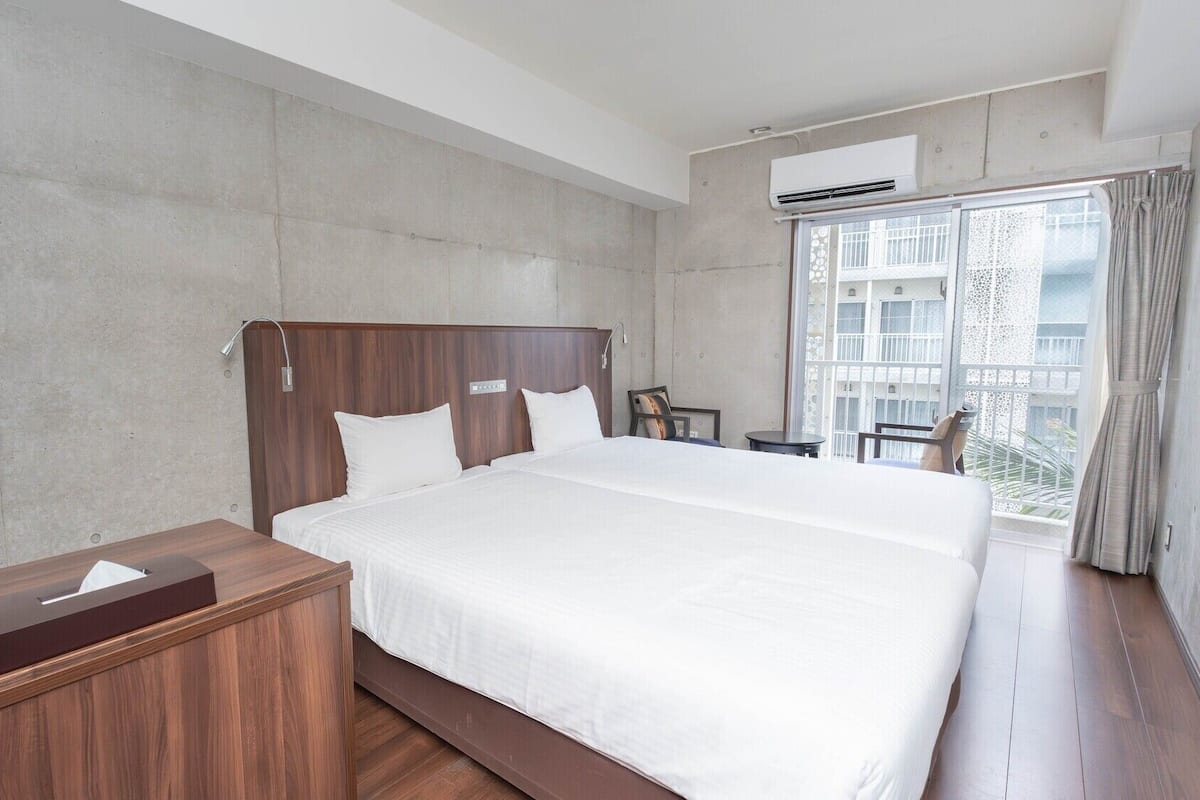 Une chambre d’hôtel avec un grand lit, une table de chevet en bois, un bureau et une fenêtre donnant sur un immeuble.