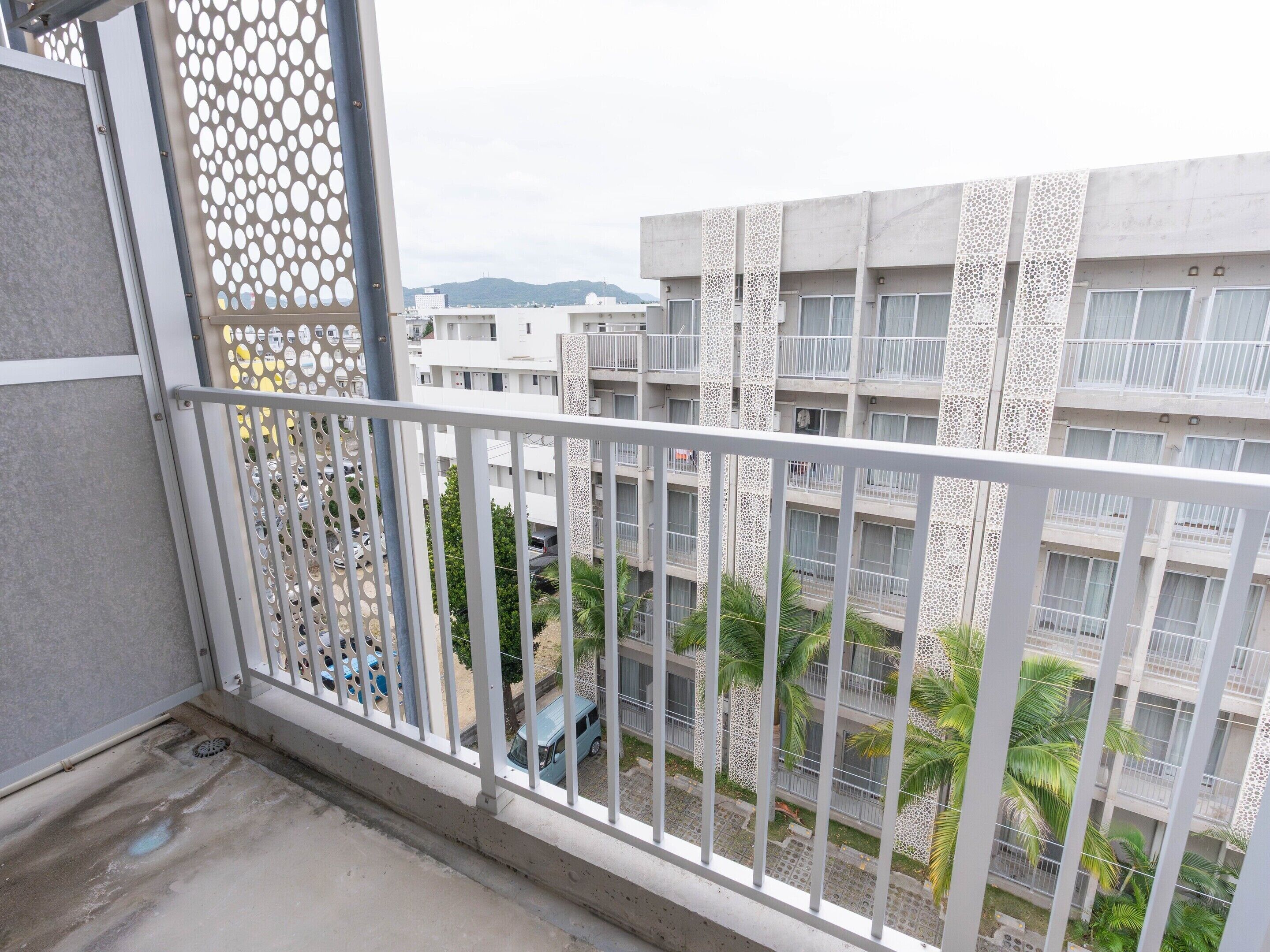 A balcony with a metal railing, a view of a multi-story building with balconies, and a palm tree in the foreground.