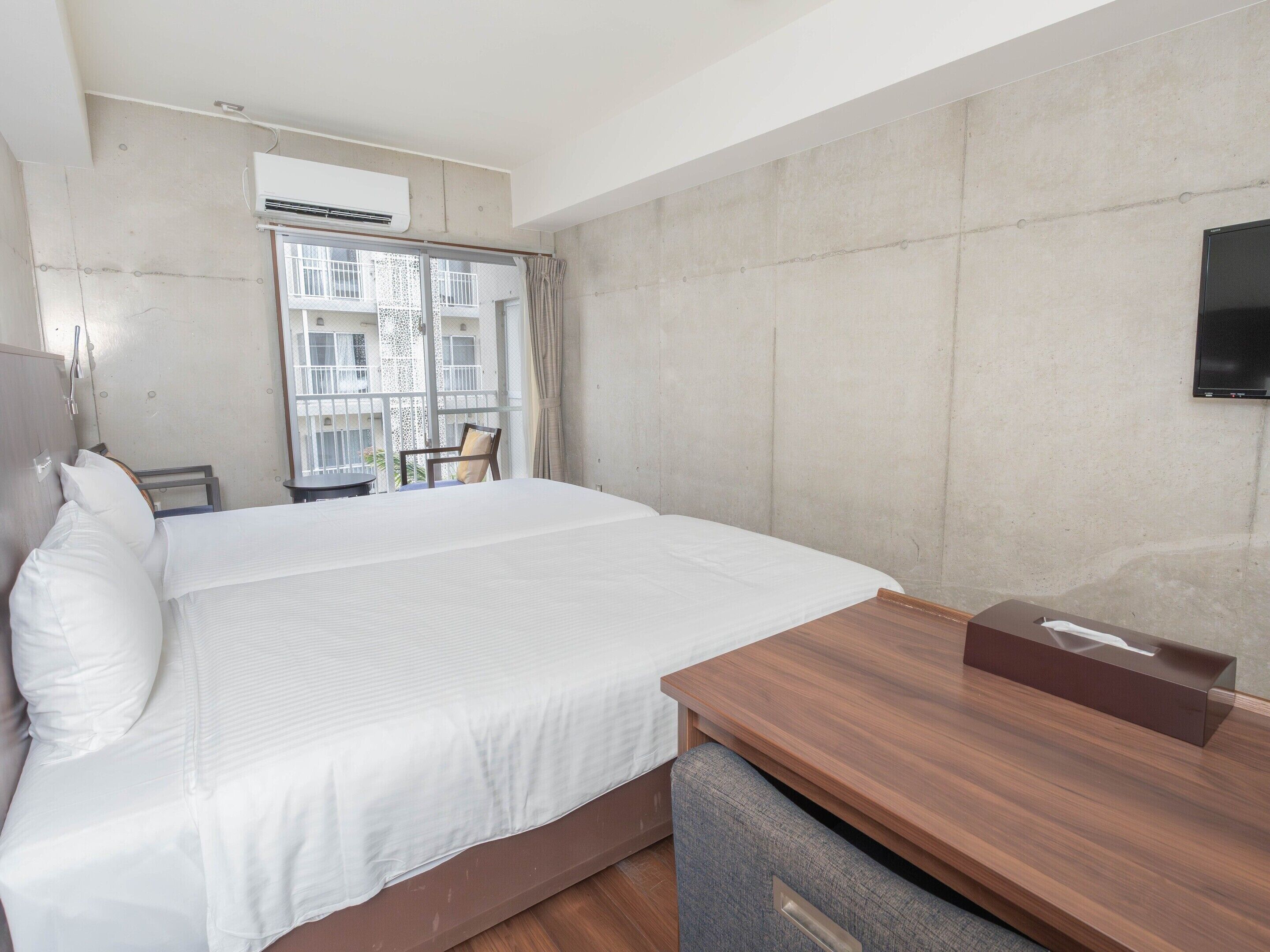 Ein Hotelzimmer mit Bett, Schreibtisch aus Holz, Fernseher und Fenster mit Blick auf ein Gebäude.