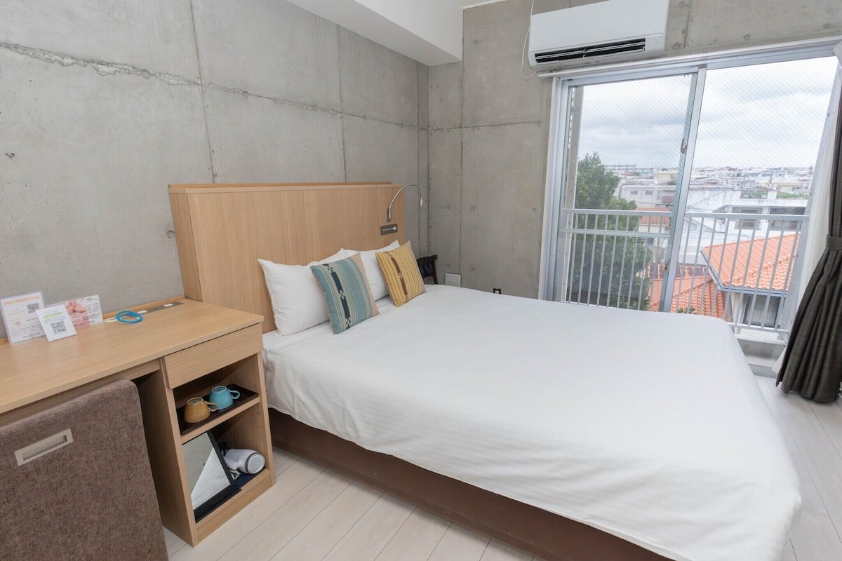 A hotel room with a bed, a desk, a small cabinet, a window with a view of buildings, and an air conditioning unit.