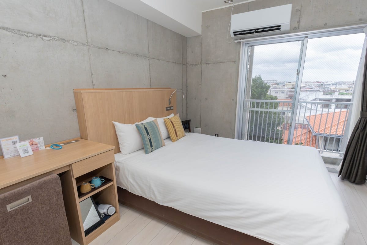 A modern hotel room with a large bed, a desk with a chair, a small wardrobe, and a window with a city view.