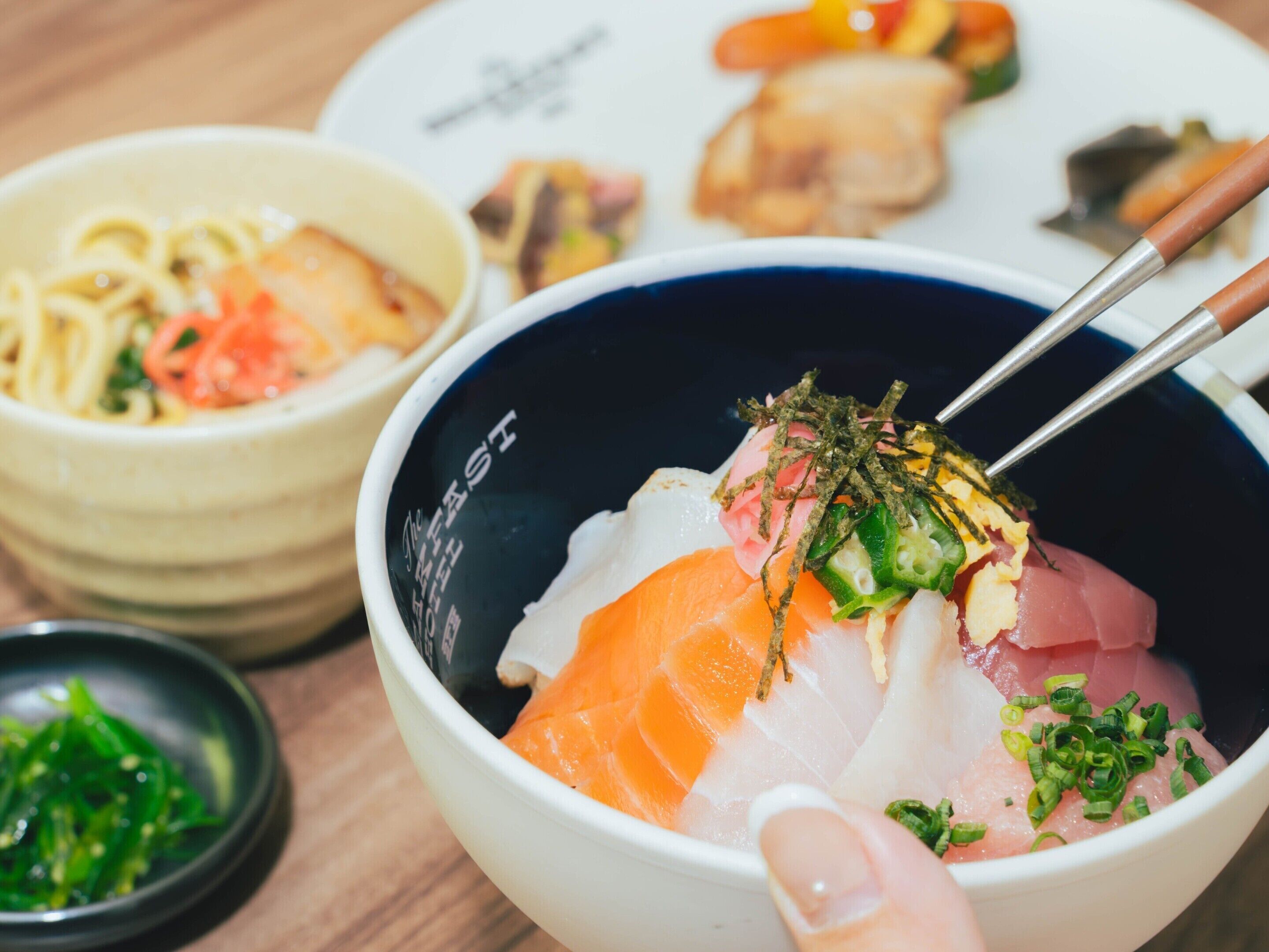 A bowl of sashimi with chopsticks, a bowl of noodles, and a plate with various dishes.