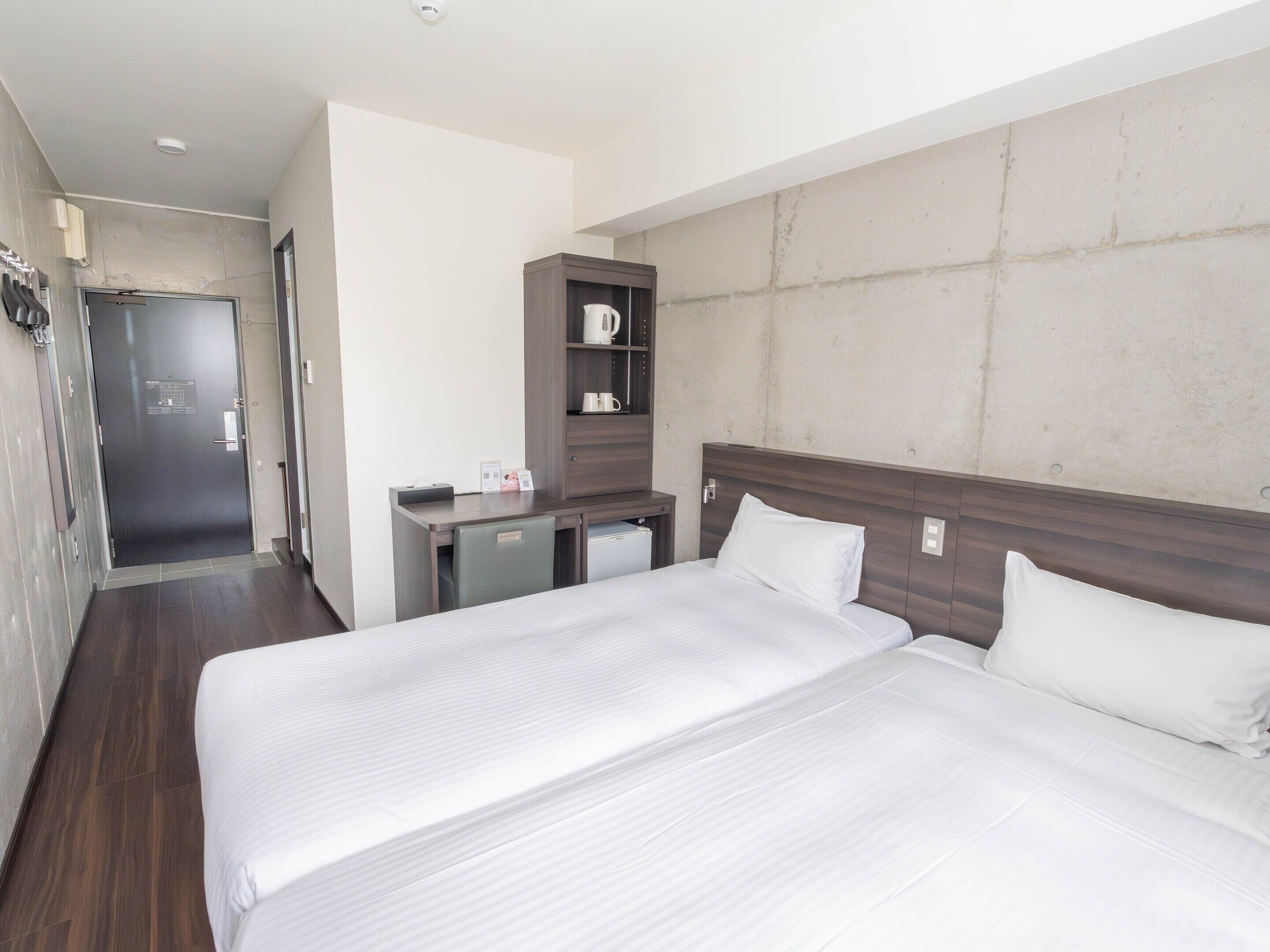 A hotel room with two beds, a desk, a small kitchenette, and a door leading to another room.