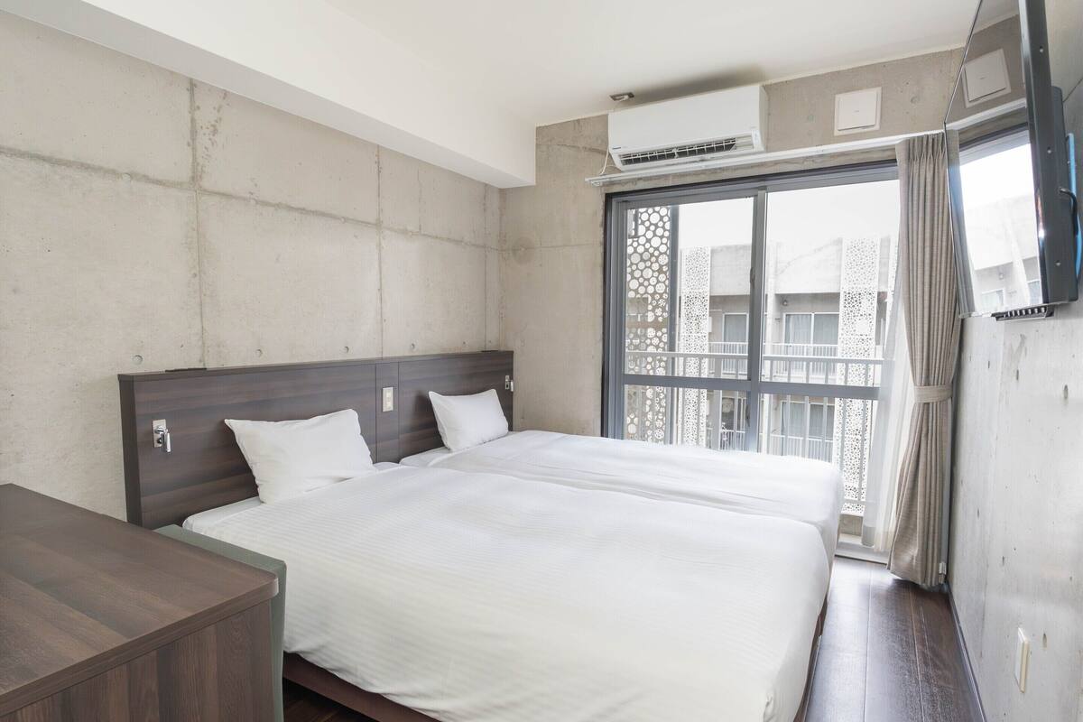 A hotel room with a large bed, a wooden desk, a window with a view of buildings, and an air conditioning unit.