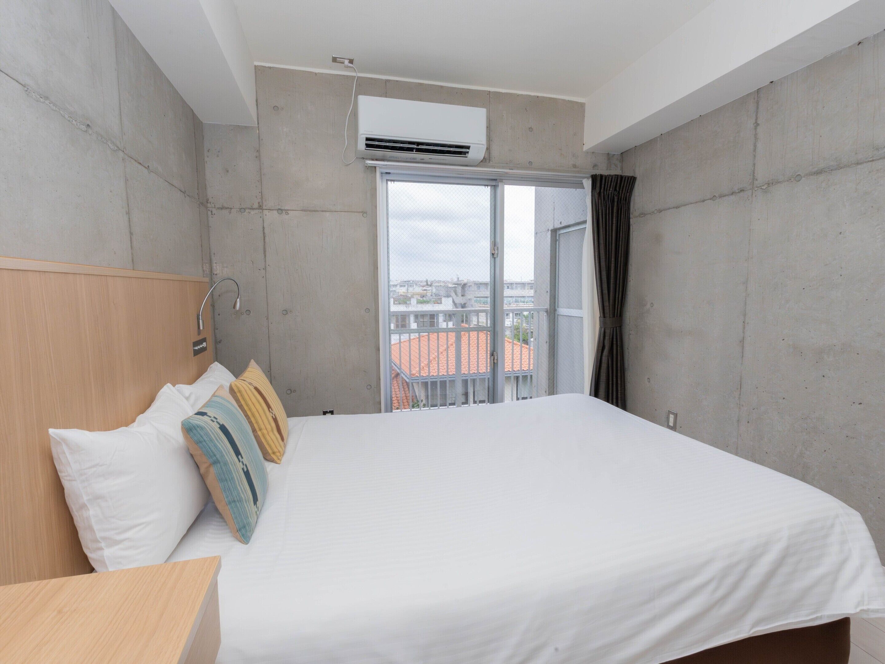 A hotel room with a large bed, a wooden headboard, a window with a view of buildings, and an air conditioning unit.