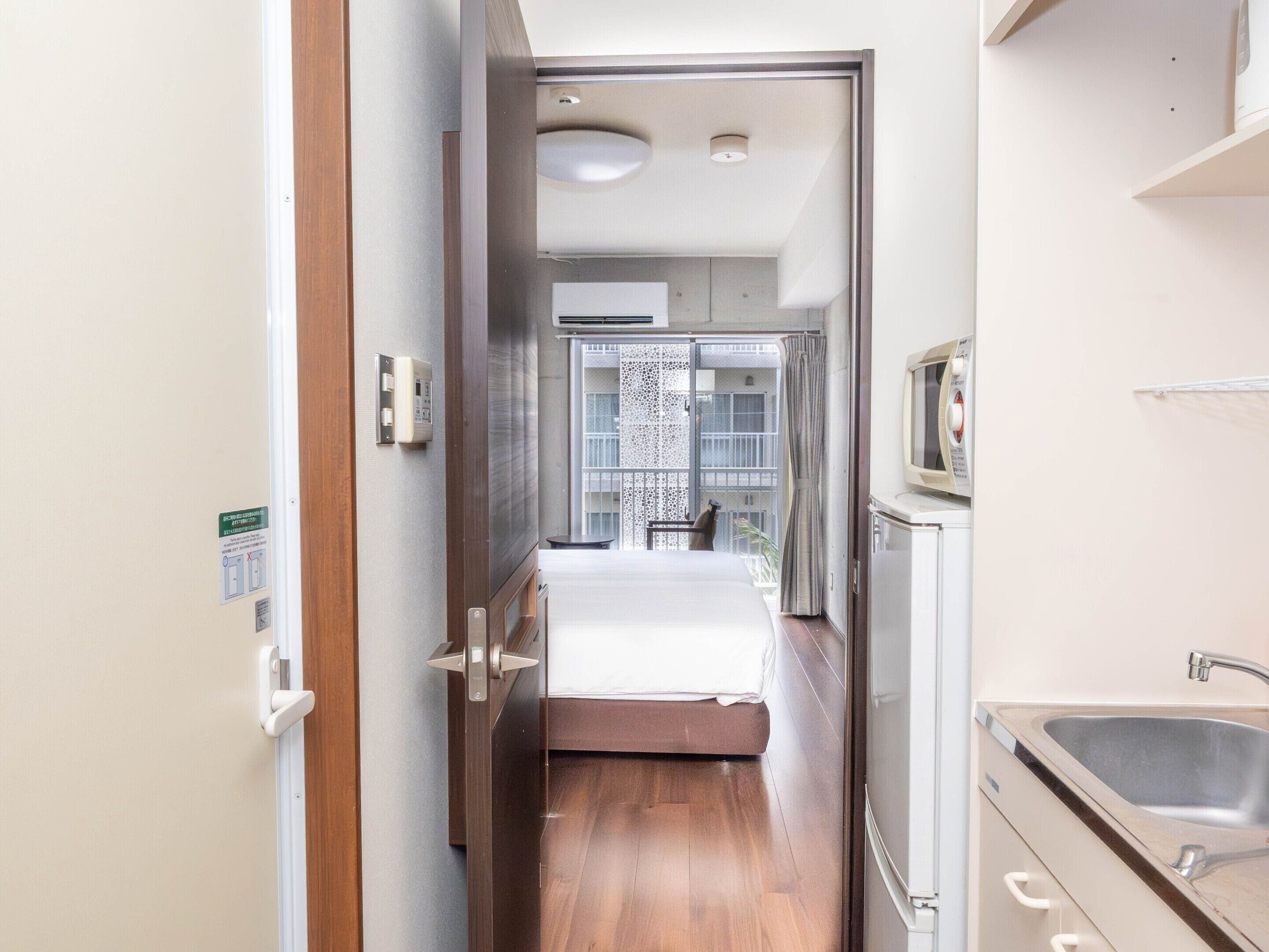 Un estrecho pasillo que conduce a un dormitorio con una cama, una mesita y una silla. Se ve una ventana con vistas a un balcón.