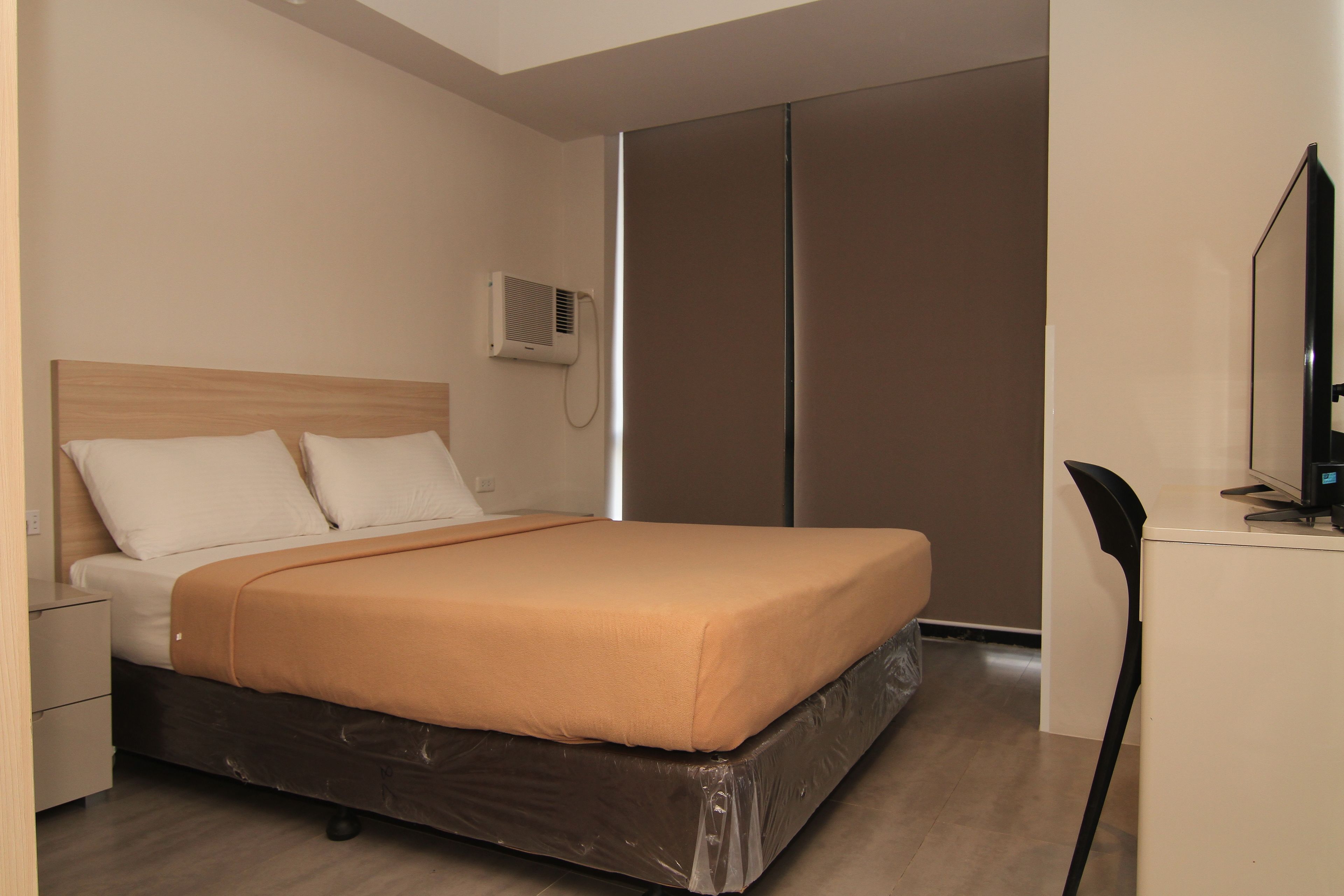 A hotel room with a bed, a nightstand, a desk with a chair, a wardrobe, and an air conditioning unit.