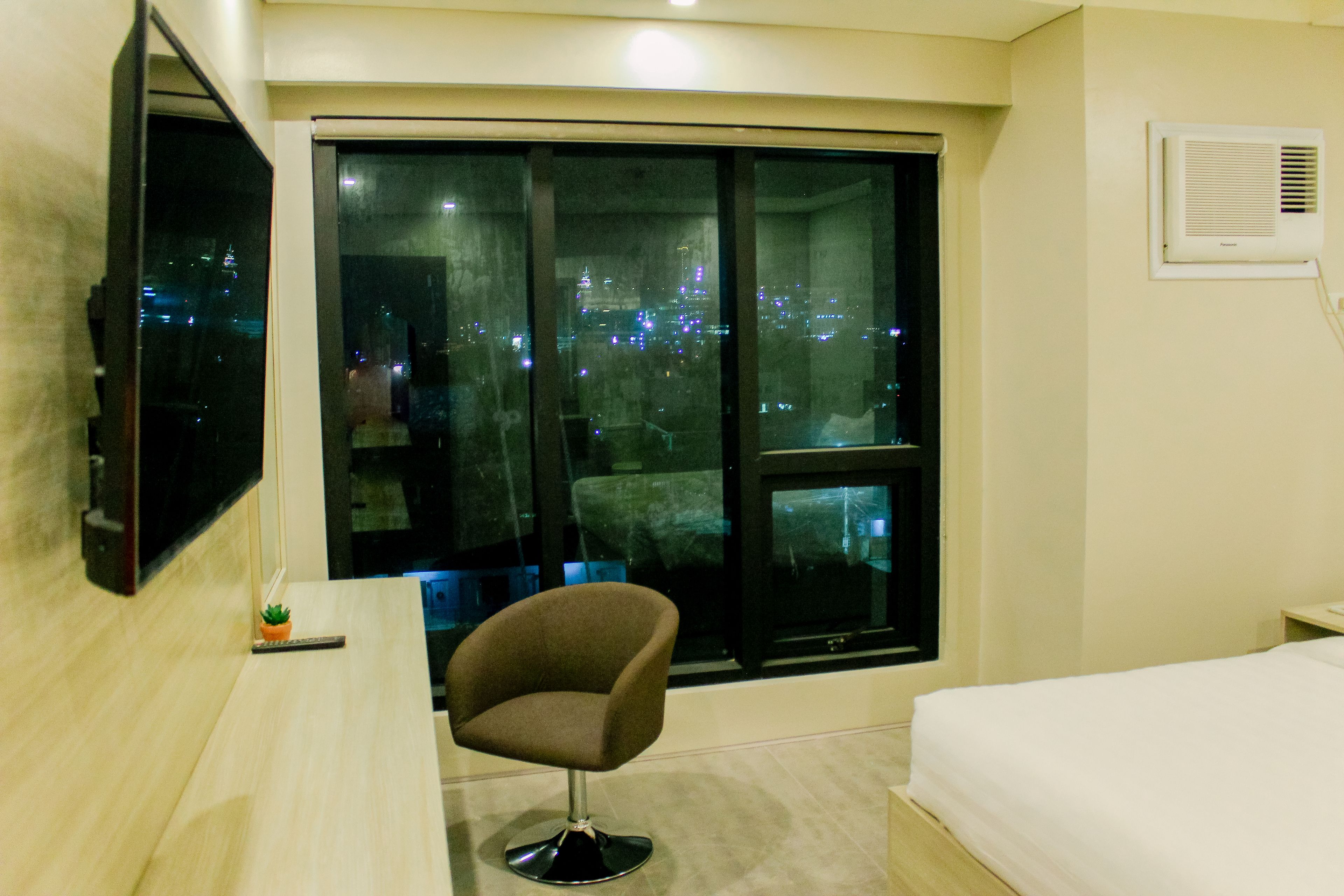 A hotel room with a large window, a flat-screen TV mounted on the wall, a small desk with a chair, and a bed.