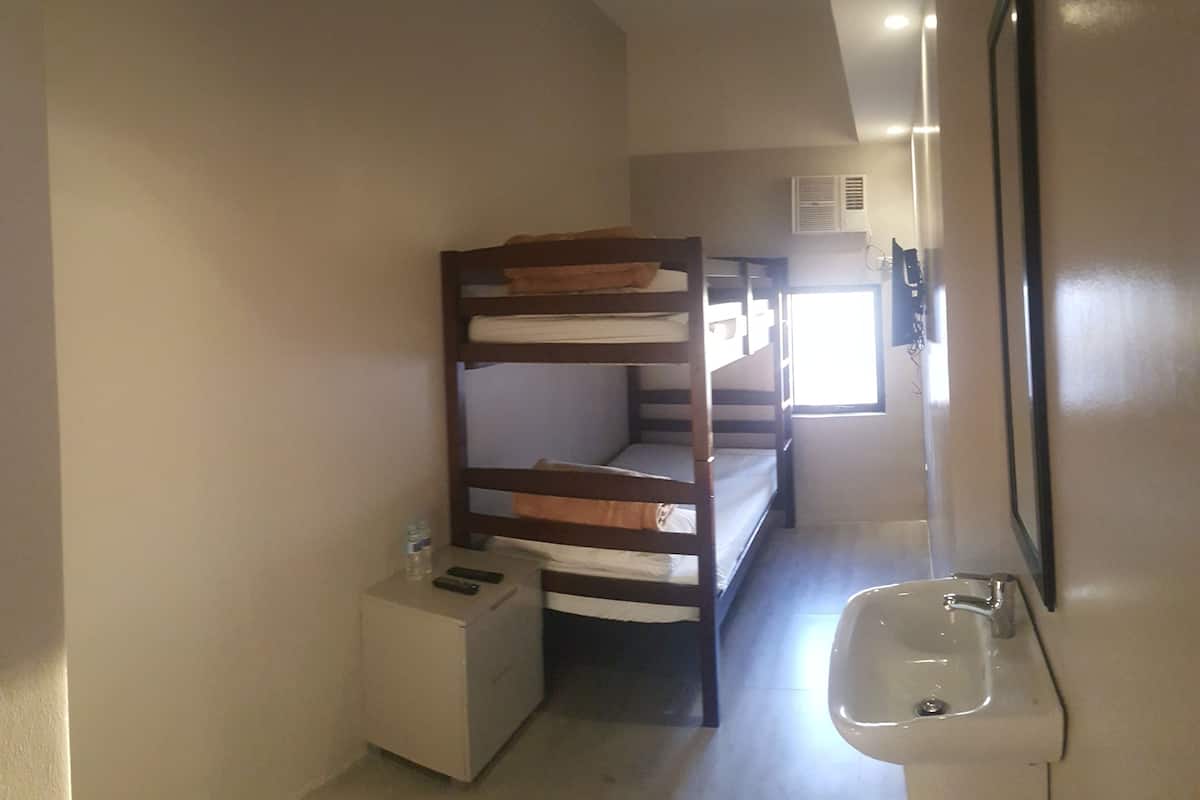 A small, compact room with a bunk bed, a small bedside table with a bottle and remote control, a sink, and a window providing natural light.