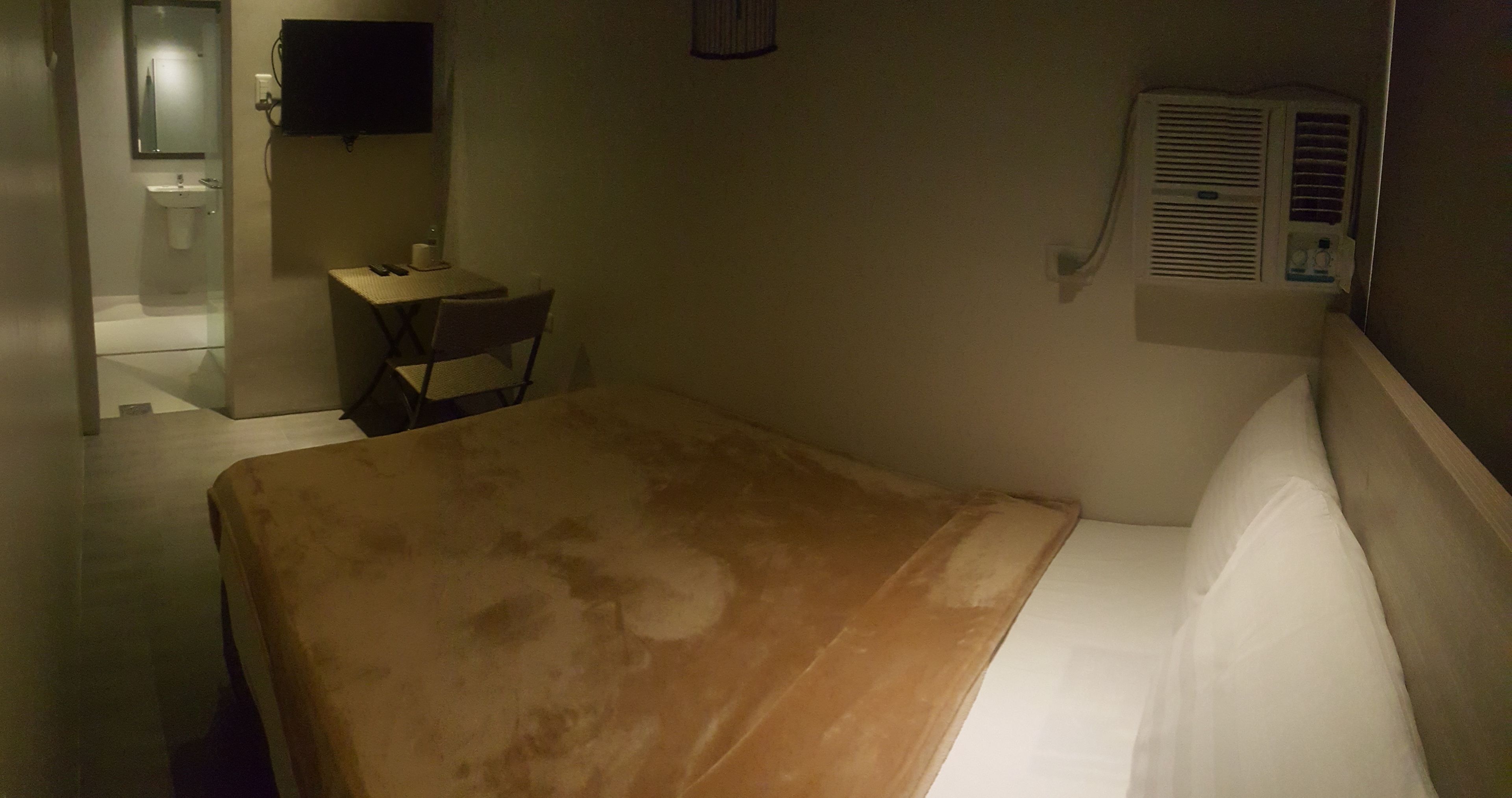 Habitación de hotel con una cama, una mesita con una silla, un televisor montado en la pared y una ventana con un equipo de aire acondicionado.