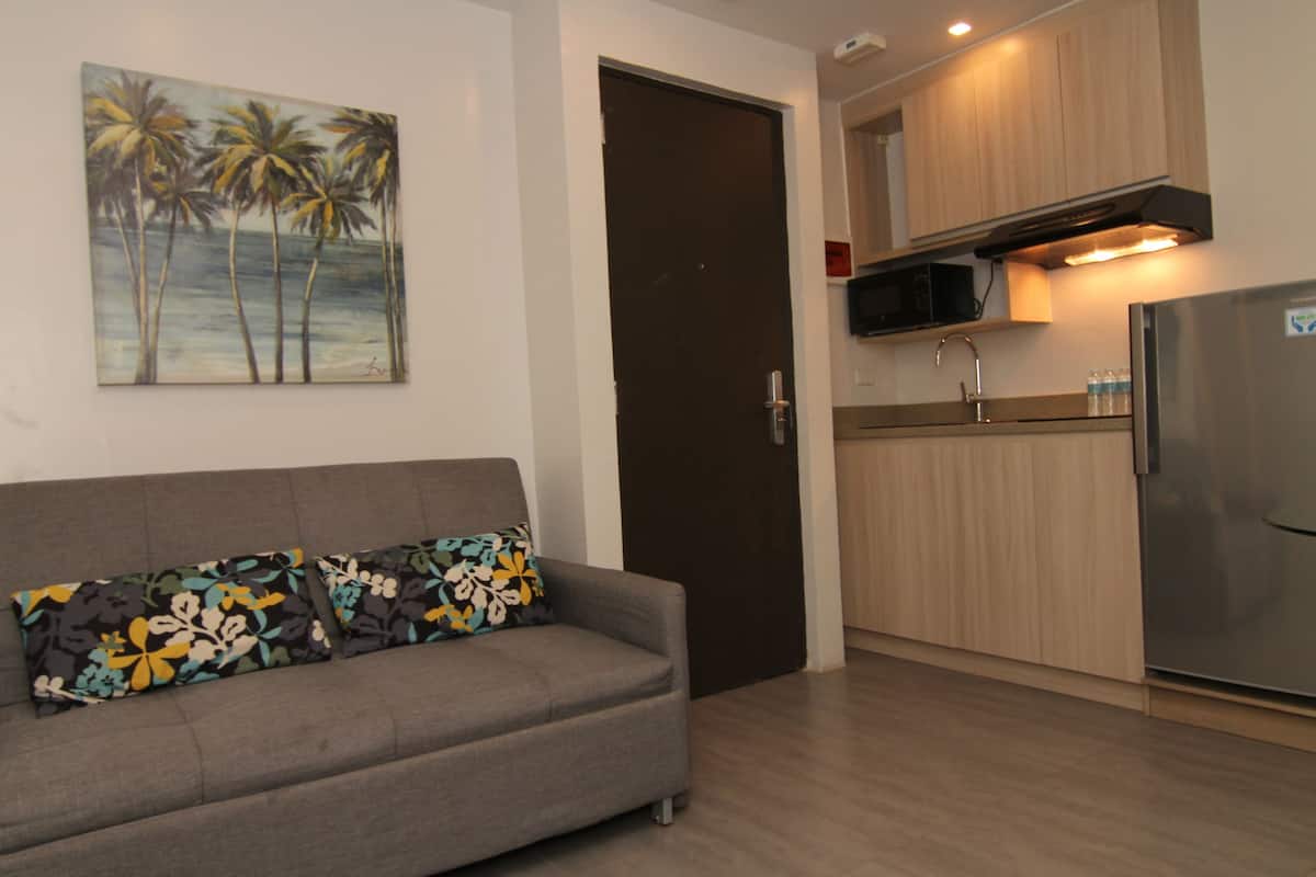 A living room with a sofa, a painting of palm trees, and a kitchen with wooden cabinets and a refrigerator.