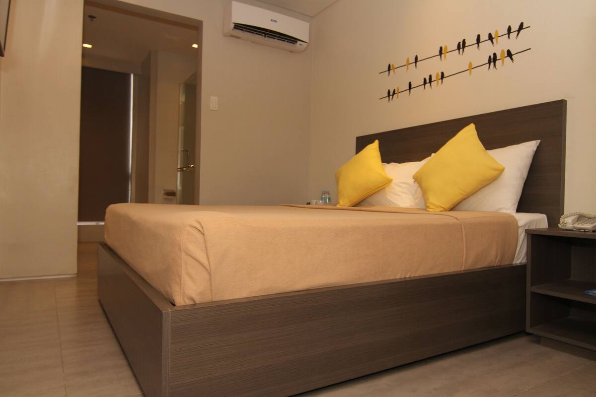 A modern hotel room with a wooden bed, beige bedding, and a wall-mounted bird art piece.
