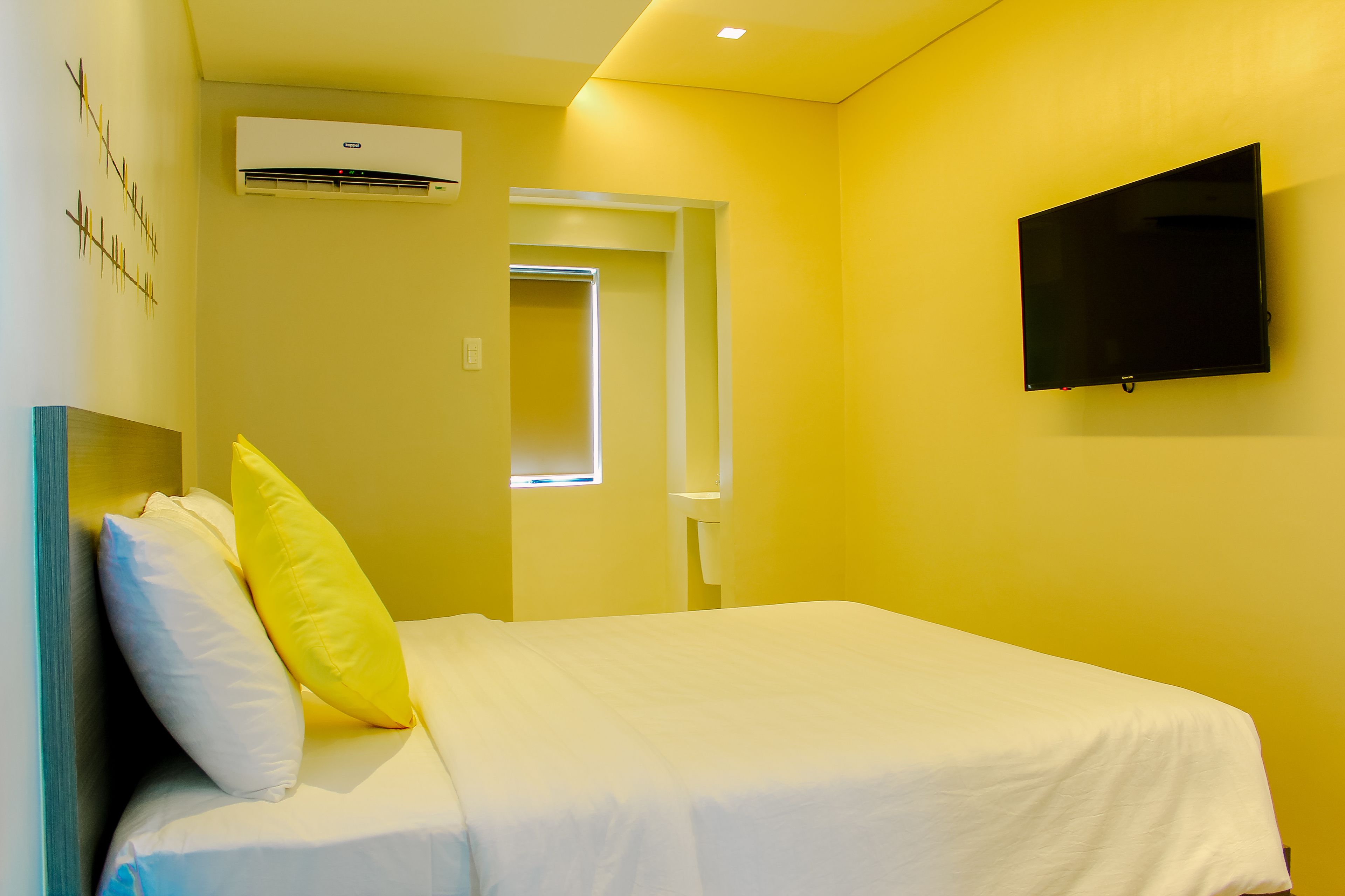 A hotel room with a bed, a flat-screen TV, an air conditioning unit, and a door leading to another room.