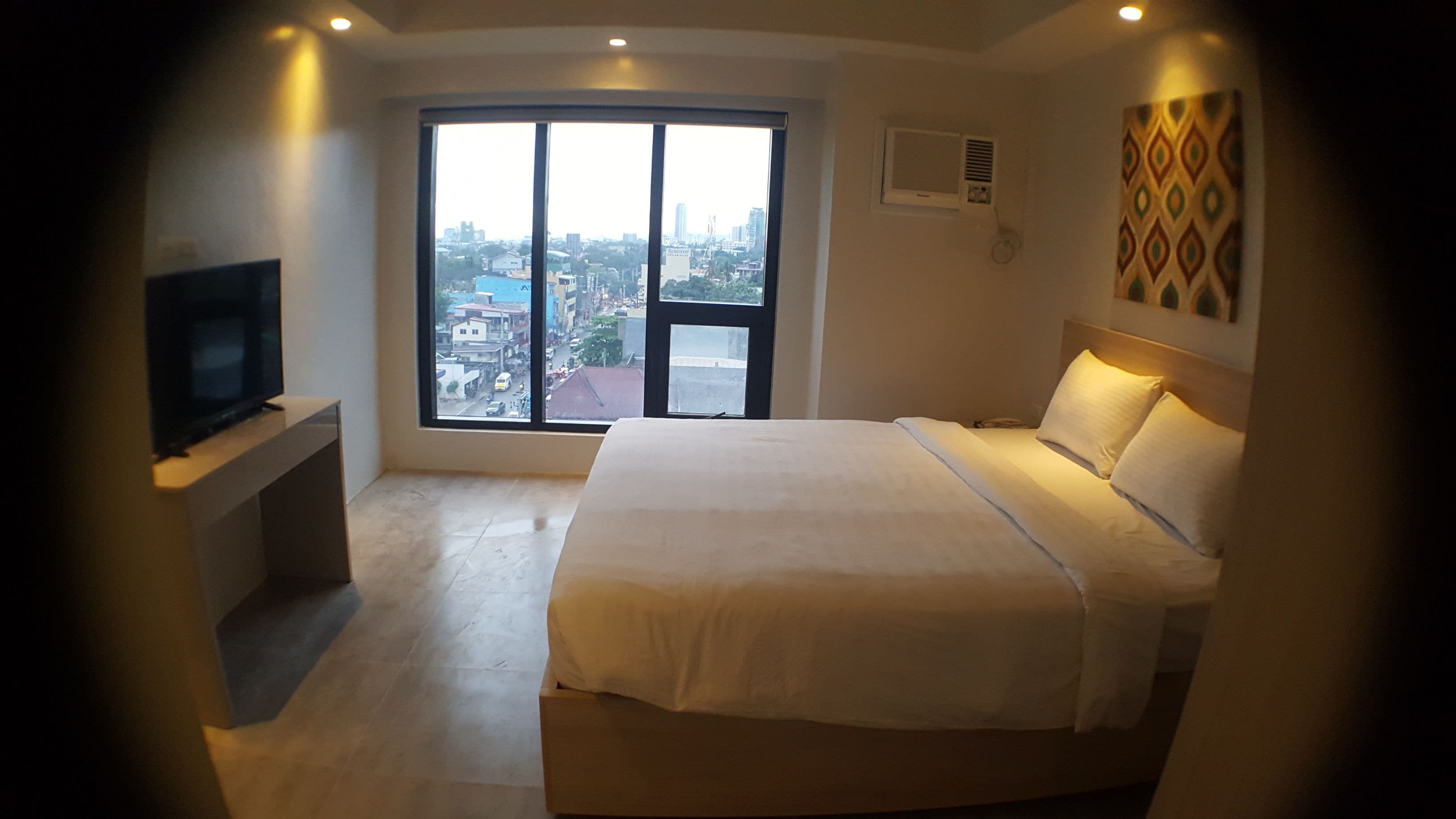 A hotel room with a large bed, a TV on a stand, a window with a city view, and a patterned wall decoration.