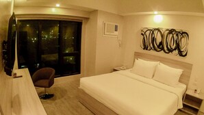 Habitación de hotel con cama, silla, escritorio, televisor y ventana con vista a la ciudad de noche.