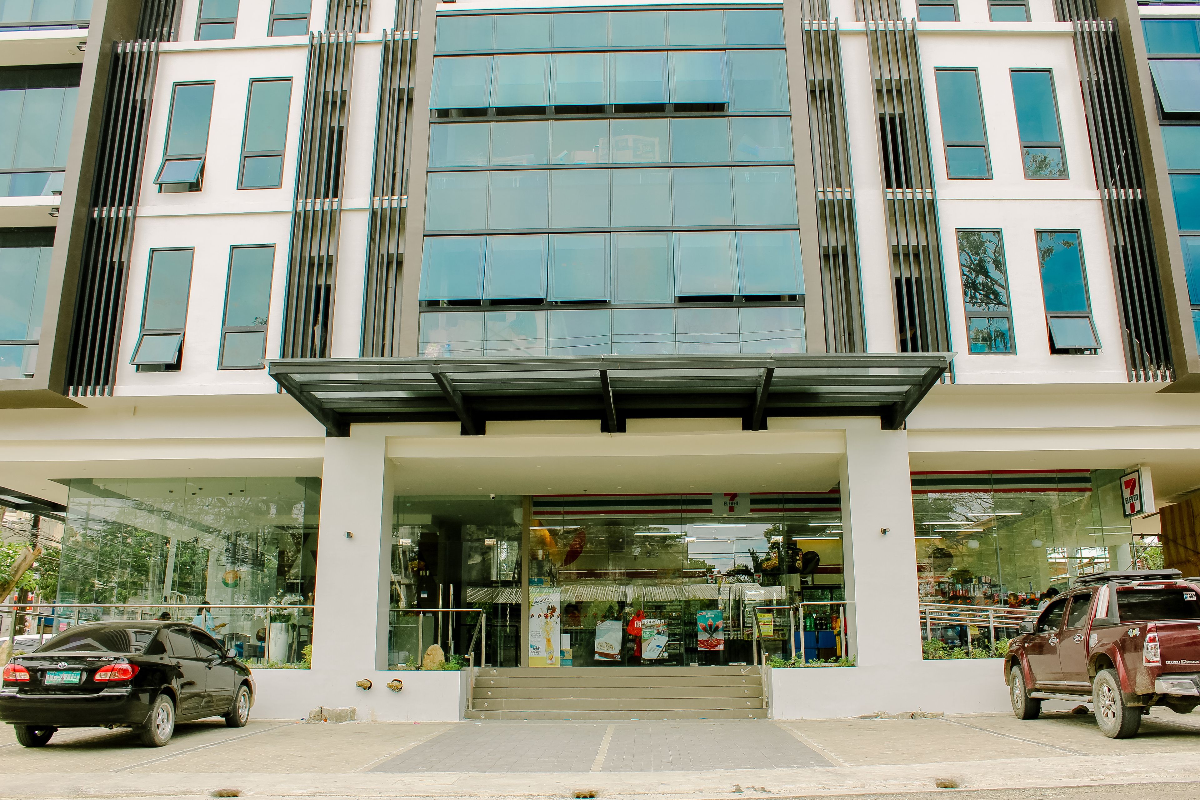 A modern building with a glass facade, a covered entrance, and a parking area with two cars.