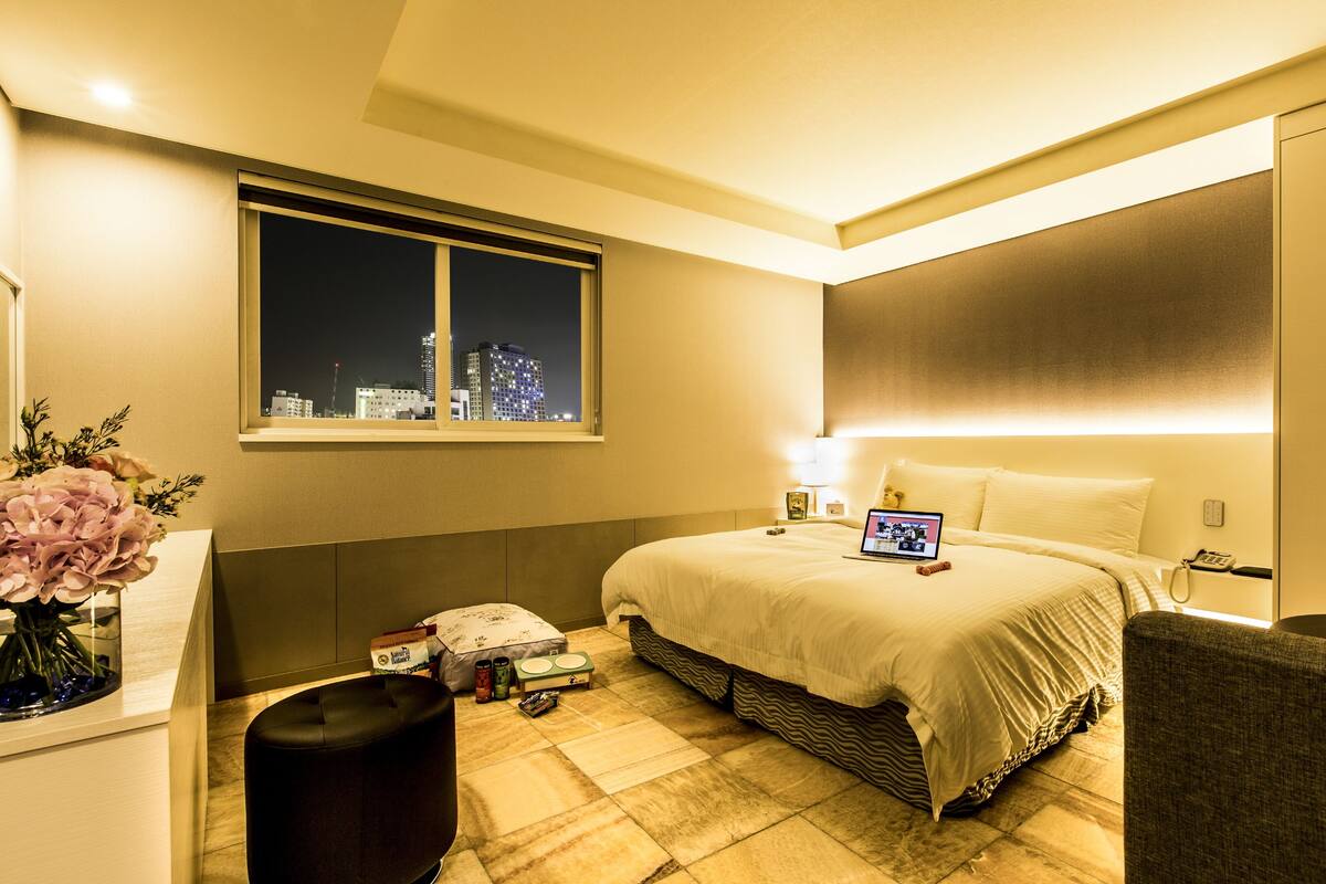 A modern hotel room with a large bed, a desk with a laptop, a nightstand, and a window with a city view.