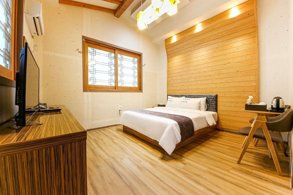 A hotel room with a wooden floor, a bed with white bedding, a desk with a computer, a chair, and a window with blinds.