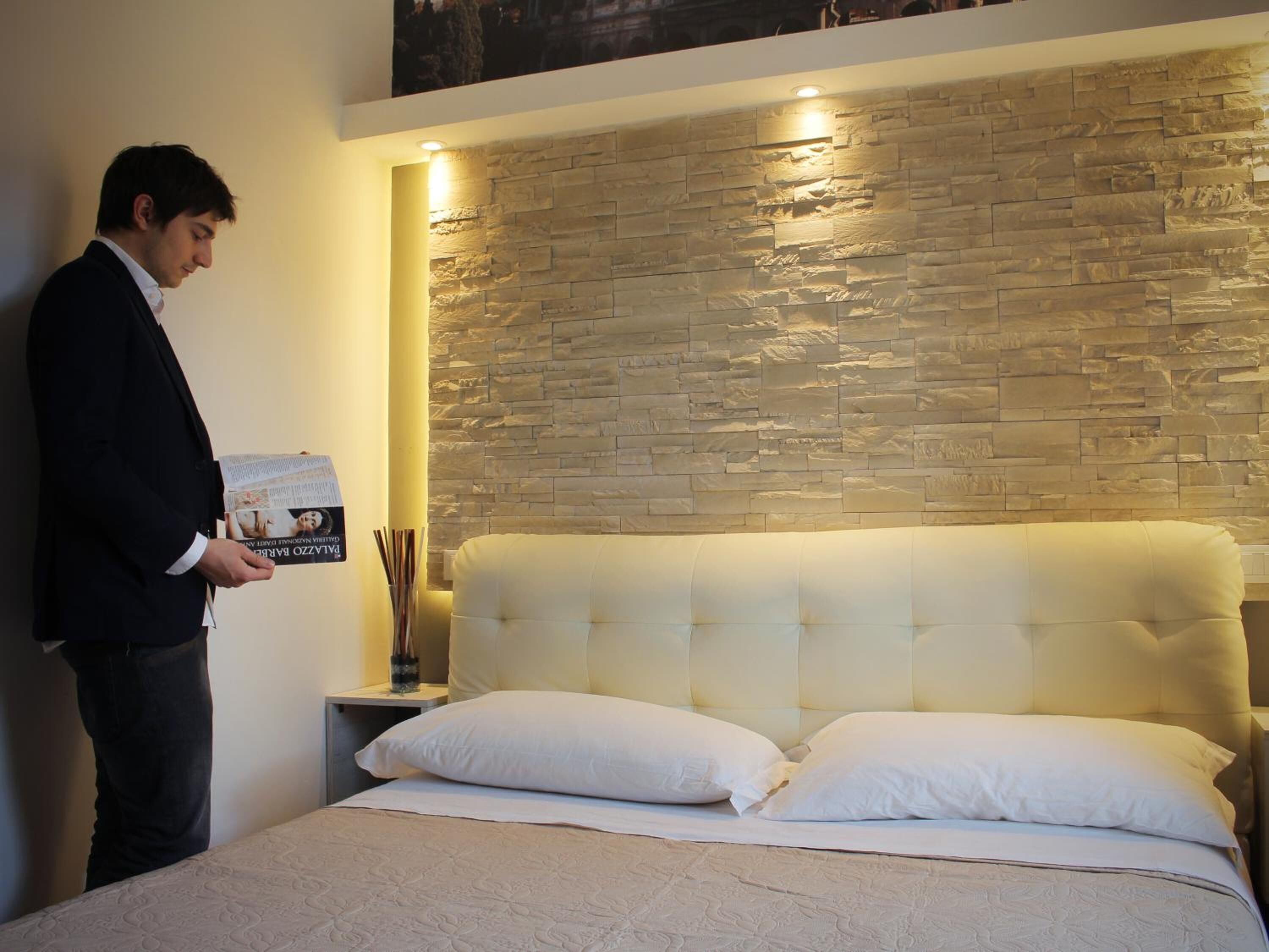 A hotel room with a stone wall headboard, a bed with white bedding, and a nightstand with a lamp and a glass of water.