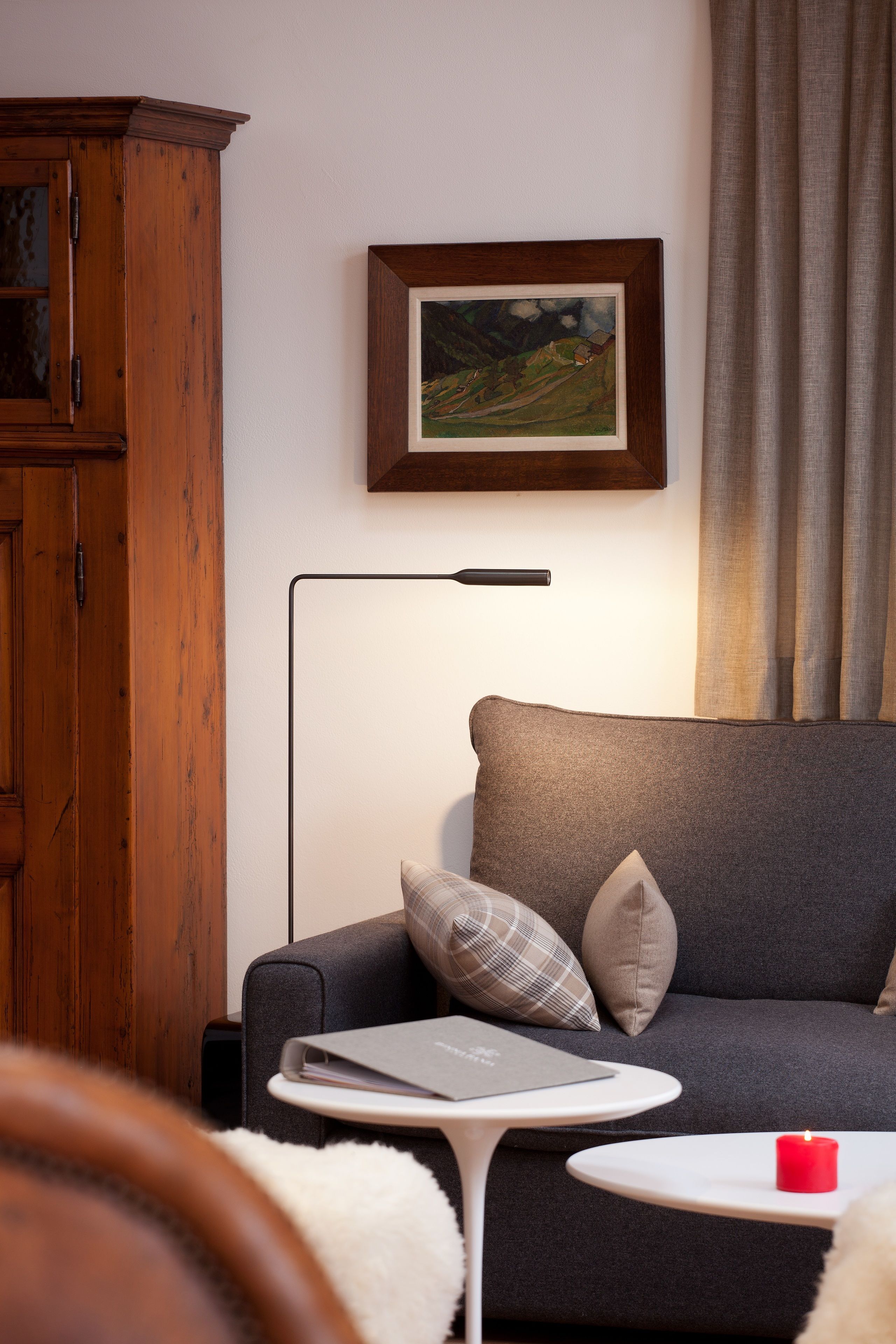A modern living room with a grey sofa, a wooden cabinet, a framed landscape painting, a white side table with a red candle, and a floor lamp.