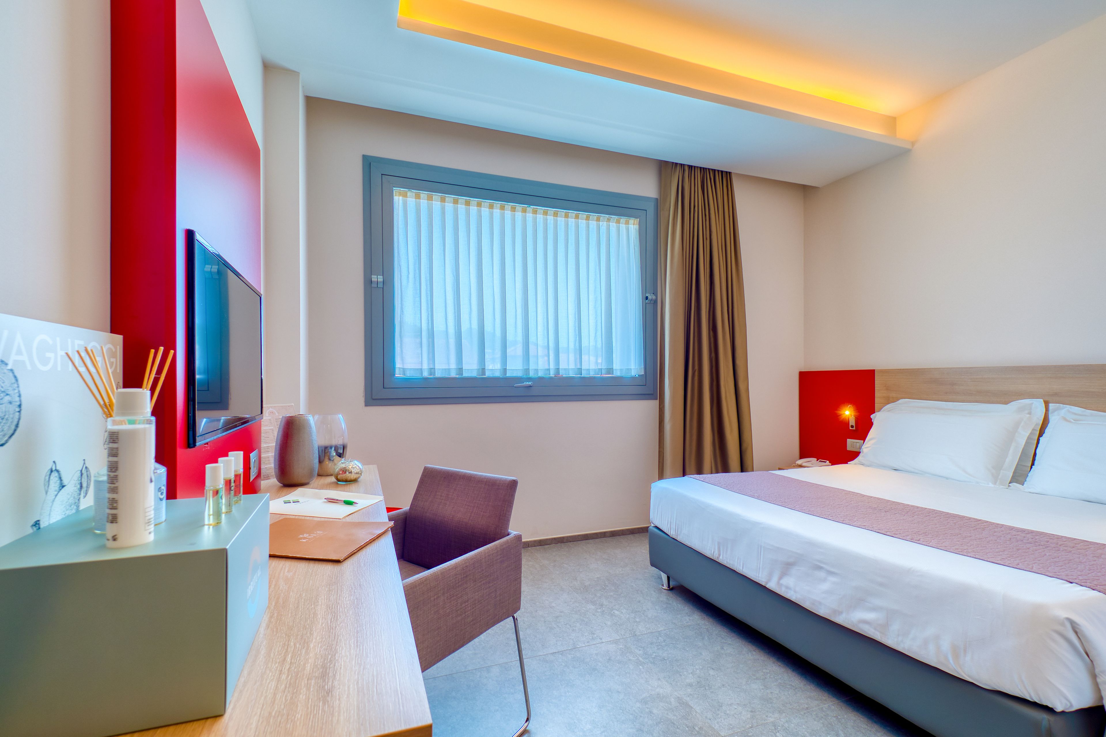 Una habitación de hotel con cama, escritorio y silla, televisor y ventana con cortinas.