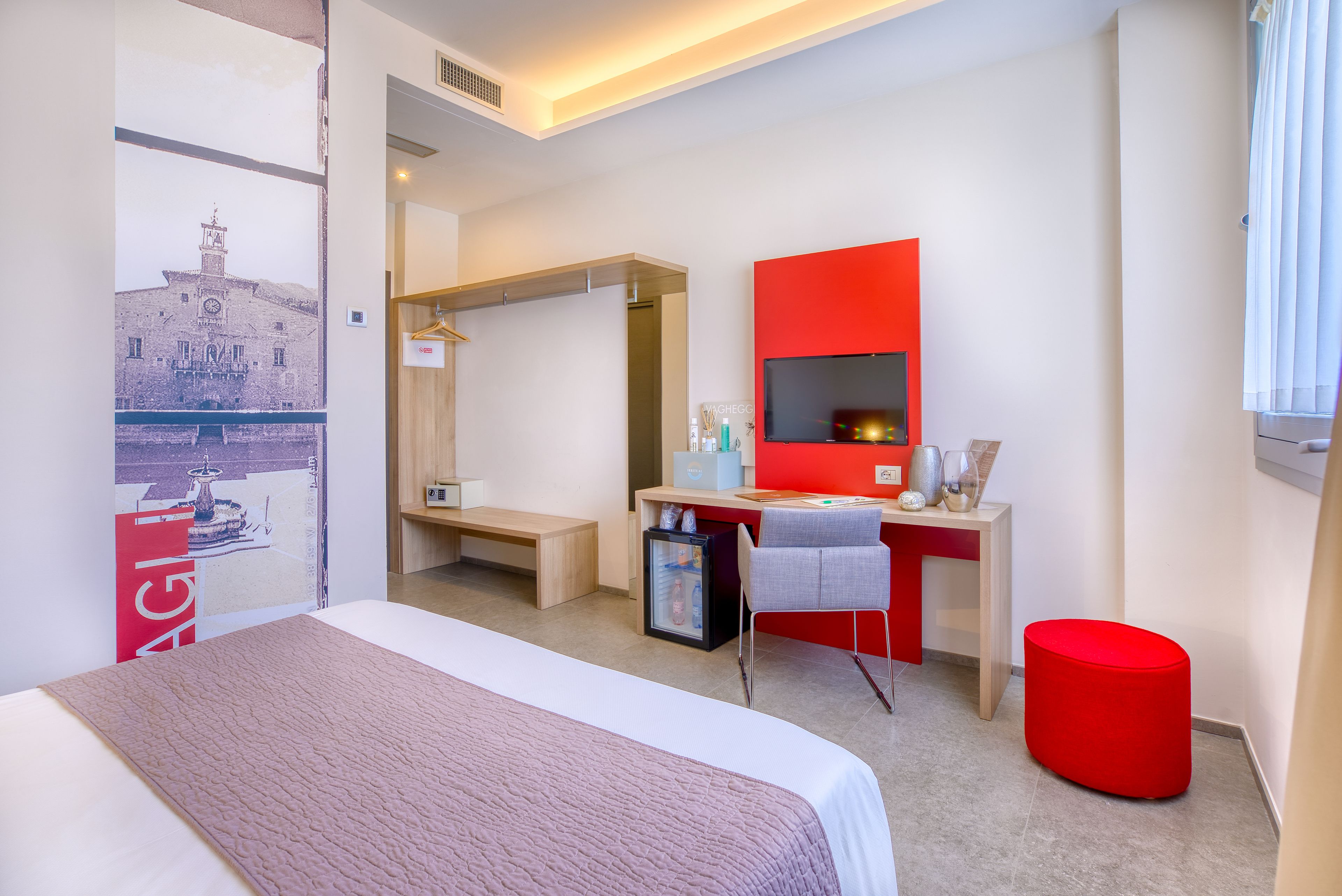A hotel room with a bed, a desk with a computer, a small fridge, a red ottoman, and a large window with curtains.