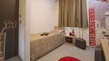 Habitación de hotel pequeña con una cama individual, una mesita de noche con lámpara, una maleta y una silla roja.