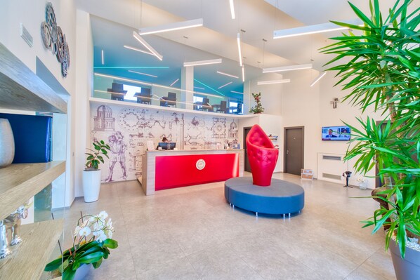 Un área de recepción moderna con un escritorio rojo, una zona de estar azul y una planta grande.