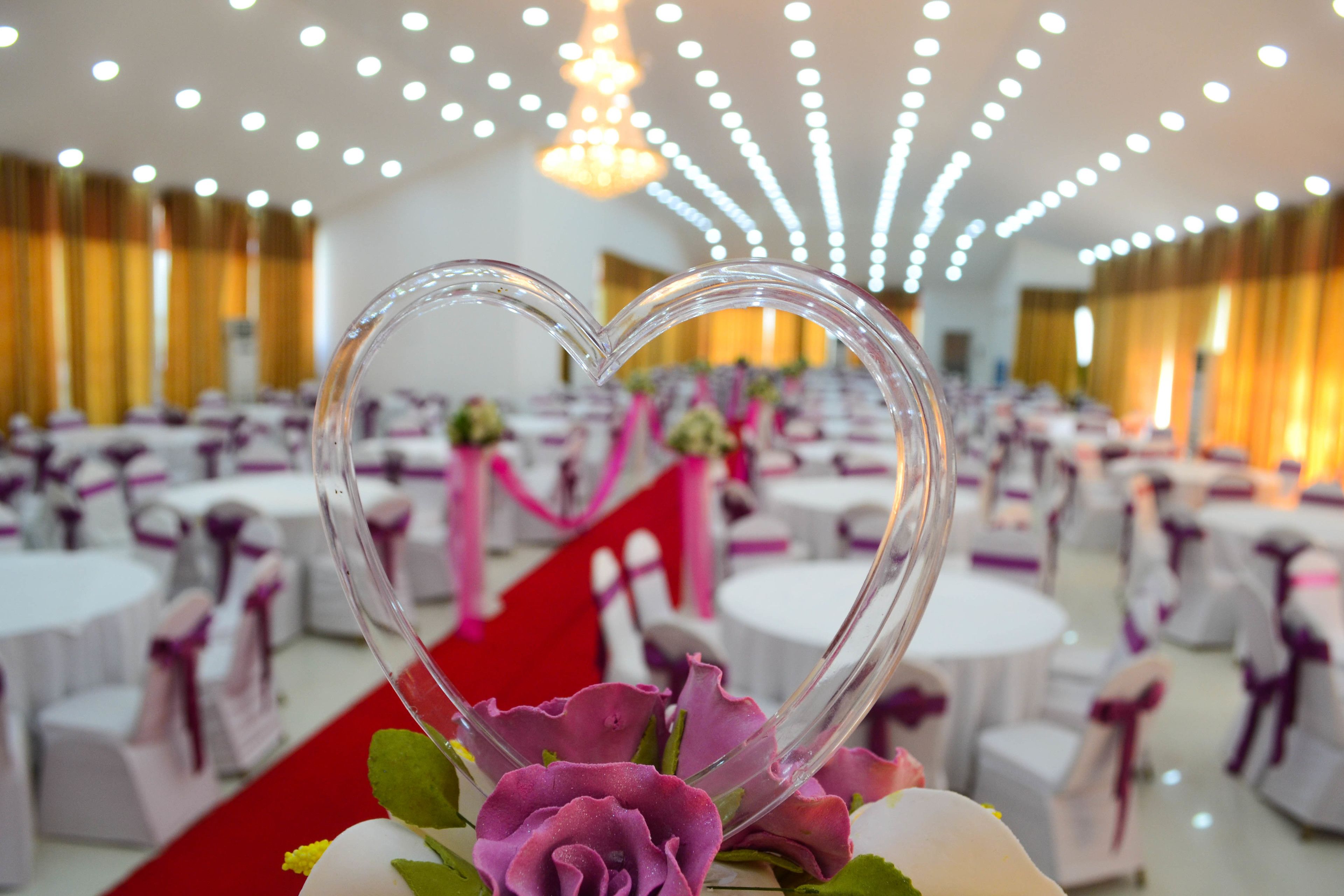 A heart-shaped decoration with flowers, set against a backdrop of round tables with white tablecloths and purple sashes, in a well-lit banquet hall.