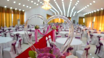 A heart-shaped decoration with flowers, set against a backdrop of round tables with white tablecloths and purple sashes, in a well-lit banquet hall.