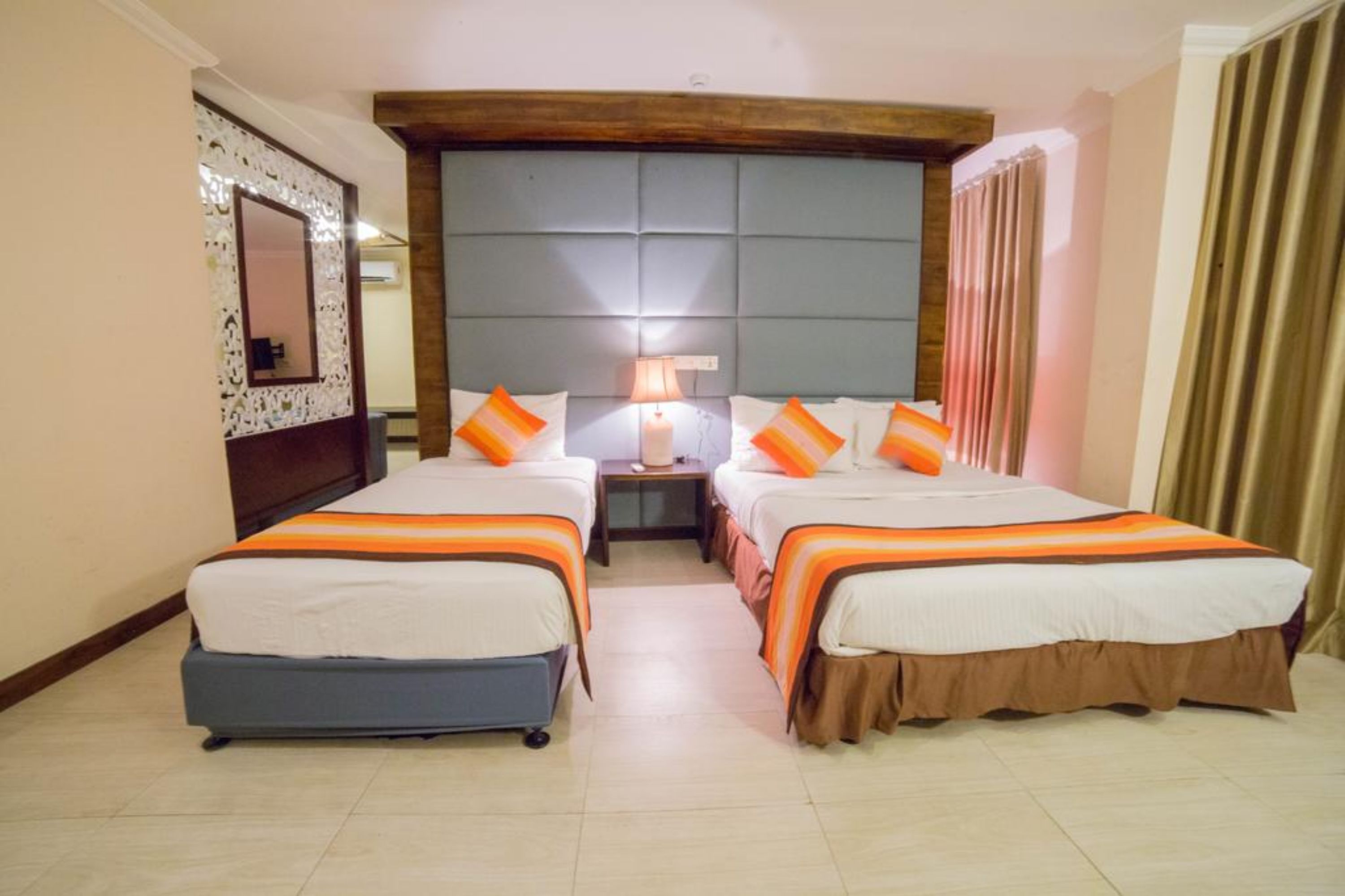 A hotel room with two beds, a nightstand with a lamp, a mirror, and a window with curtains.