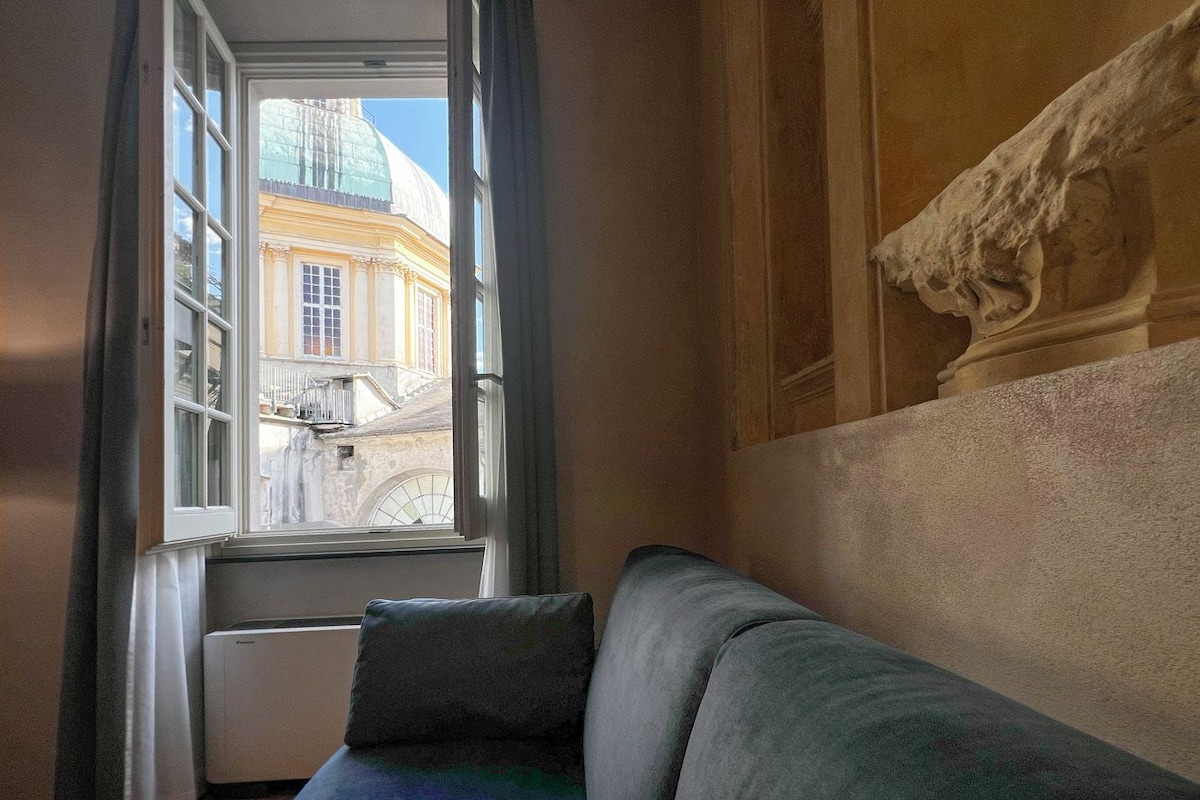 A room with a view of a building through a window, a sofa, and a radiator.
