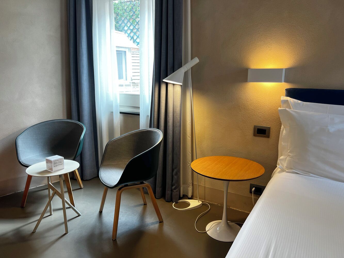 A hotel room with a bed, two chairs, a small round table, a lamp, and a window with curtains.