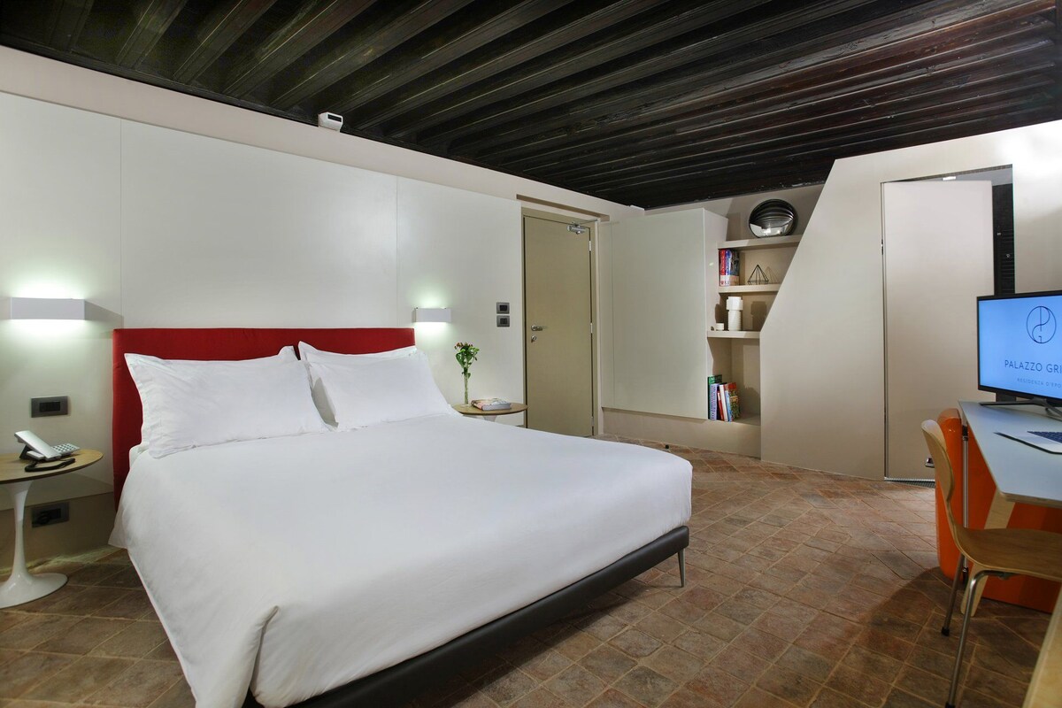 A hotel room with a bed, bedside table, lamp, and a desk with a computer.