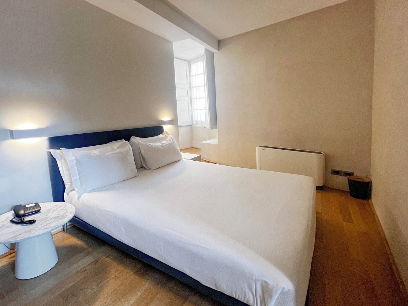 A modern hotel room with a large bed, bedside table with a lamp, and a small table with a phone.
