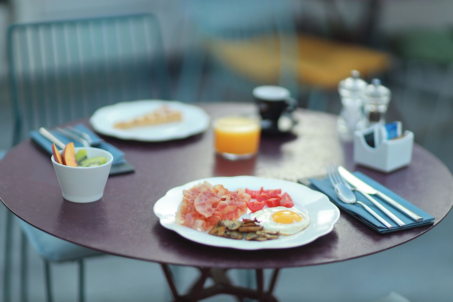 A breakfast setting with a plate of eggs, bacon, and tomatoes, a bowl of fruit, a glass of orange juice, and a cup of coffee on a round table.