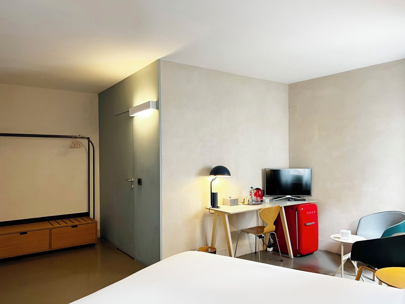 A hotel room with a bed, a desk with a TV, a red fridge, and a chair.
