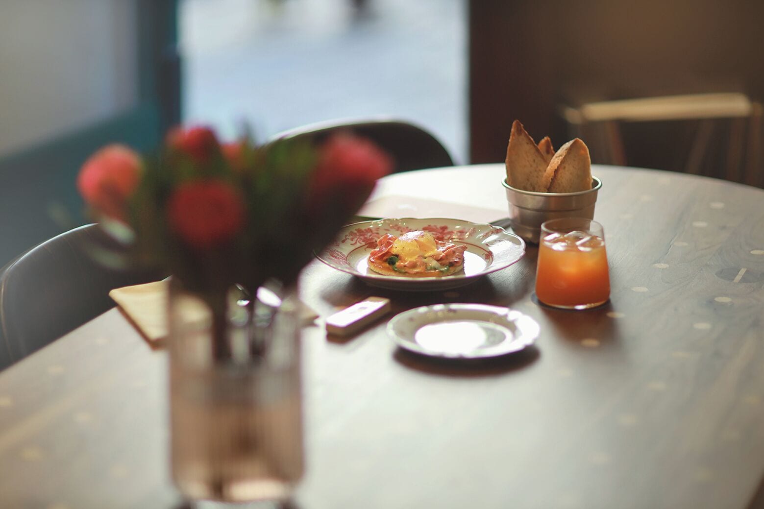 Una mesa de comedor con un plato de comida, un vaso de jugo de naranja y un pequeño jarrón con flores.