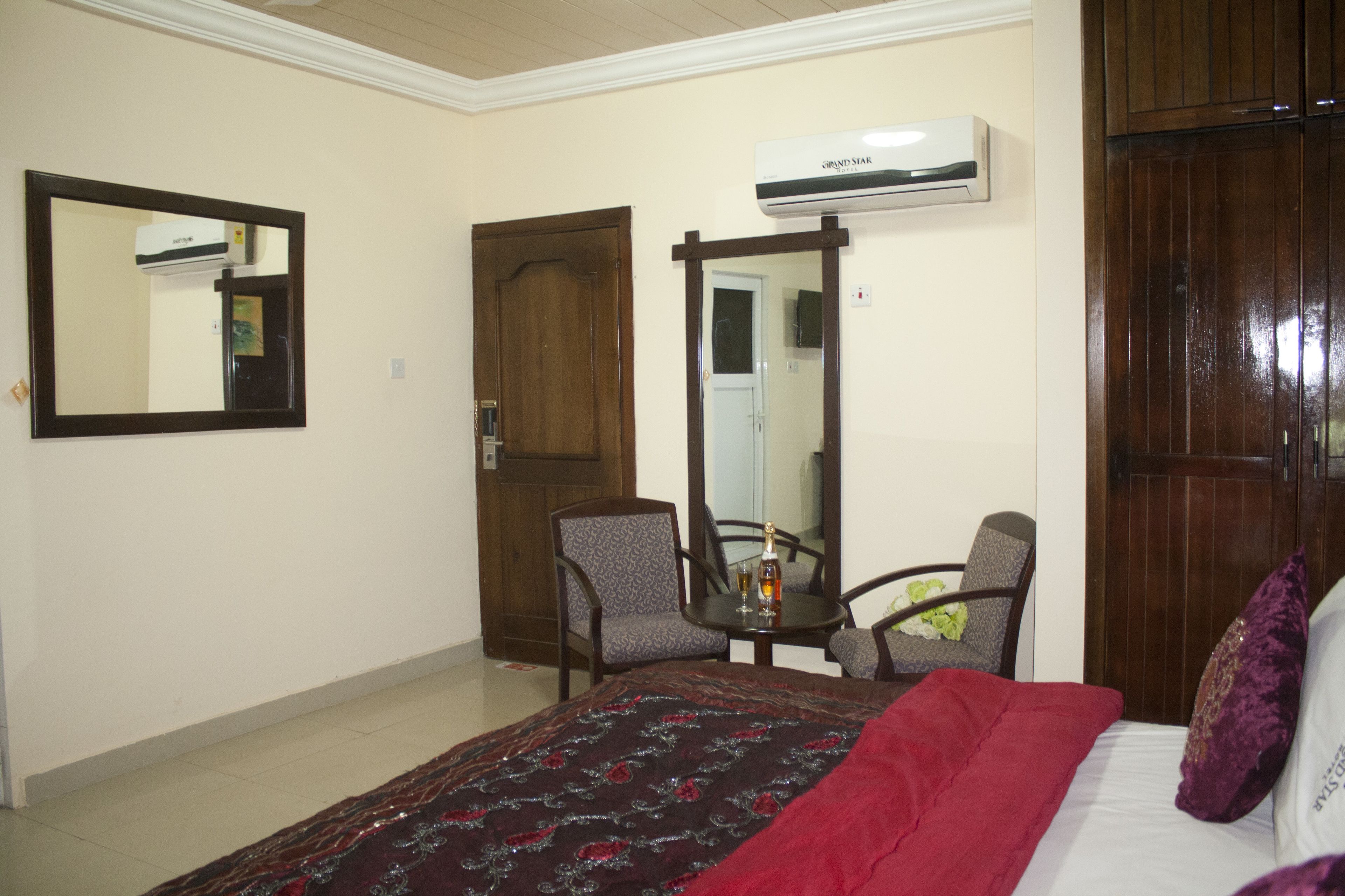 A hotel room with a bed, a wooden wardrobe, a mirror, a small table with chairs, and an air conditioning unit.