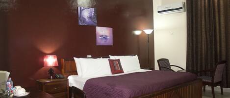 A hotel room with a large bed, a desk with a lamp, a chair, and artwork on the wall.