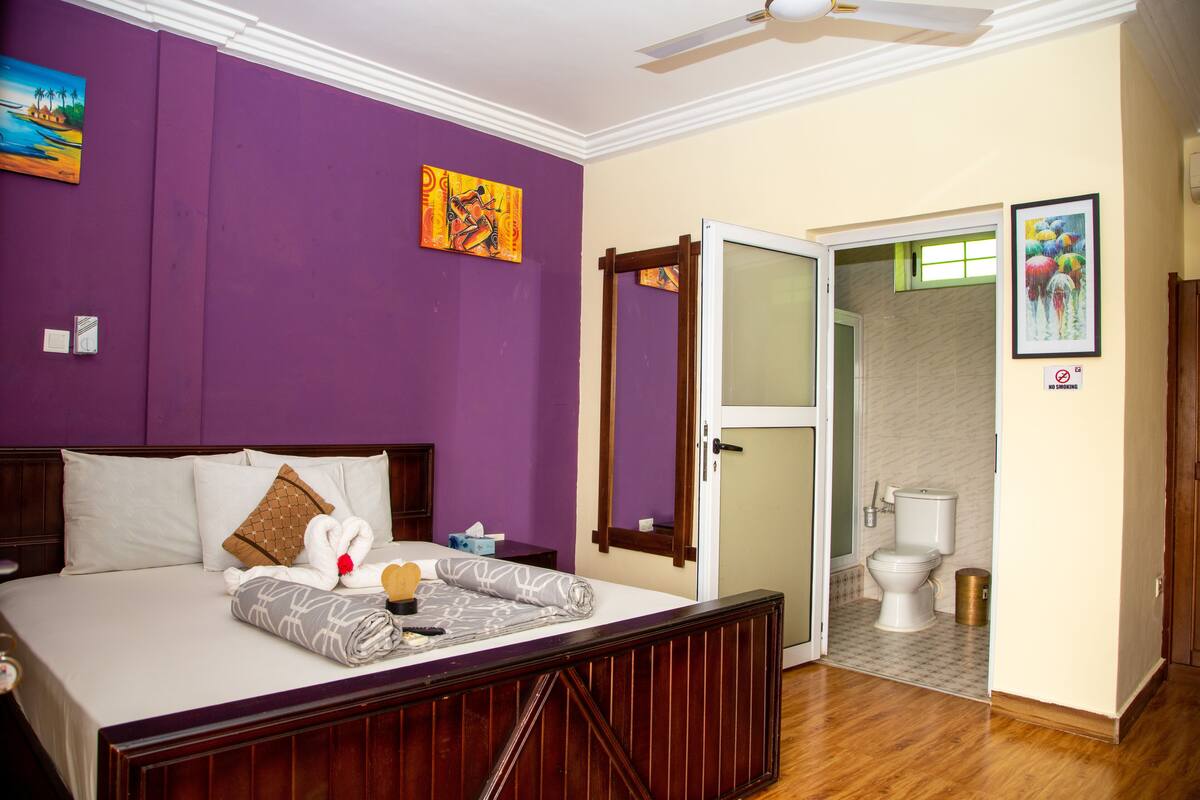 A hotel room with a bed, a wooden headboard, a ceiling fan, and a bathroom visible through an open door.