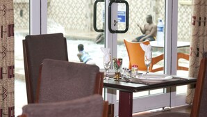 A dining area with a table set for two, a window view of a pool area, and a patterned curtain. - Grand Star Hotel (Accra)