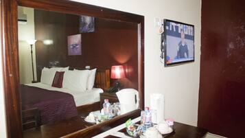 A hotel room with a large mirror, a bed with white linens, a bedside table with toiletries, and a wall-mounted television.